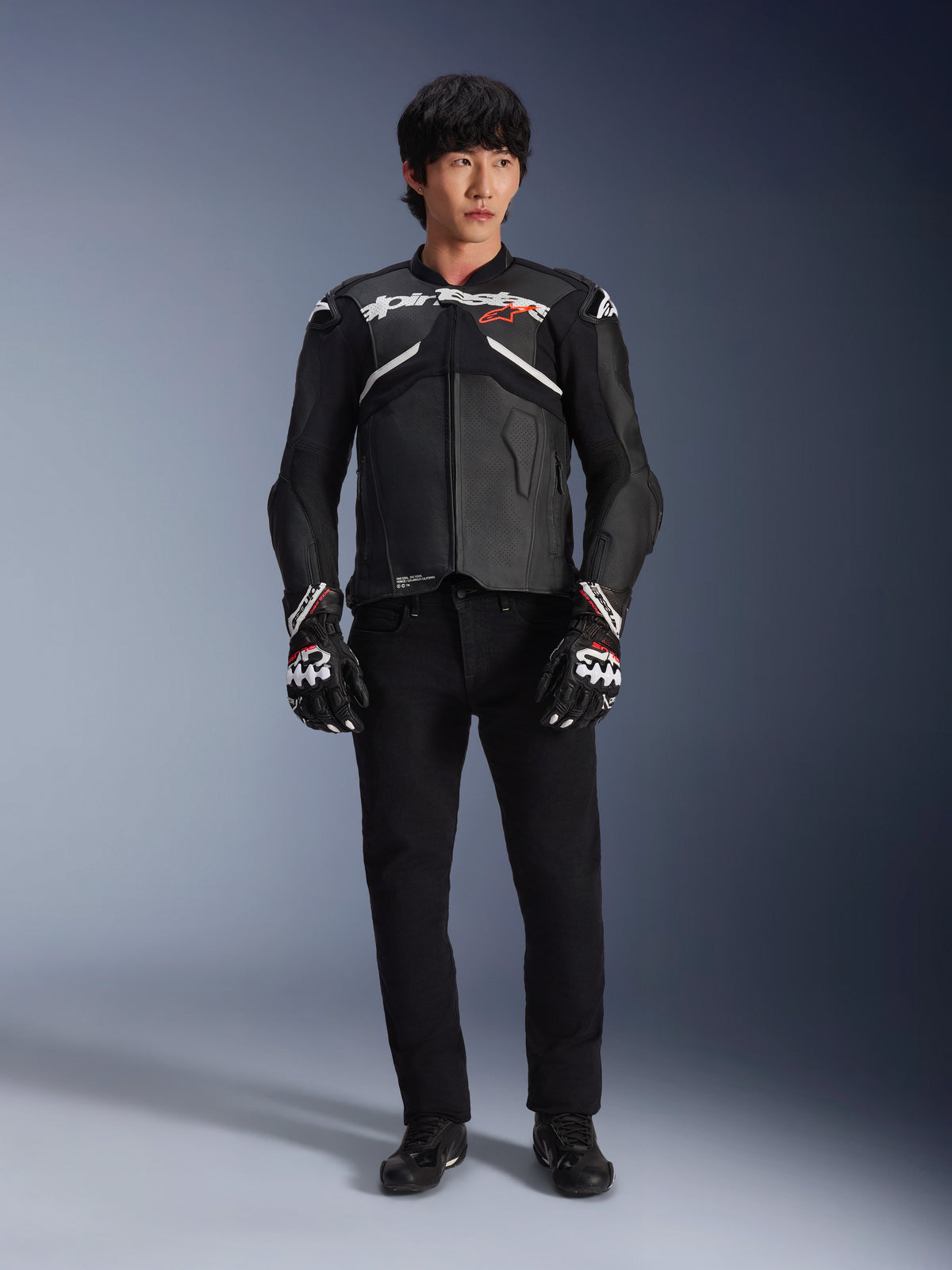 Model wearing the Alpinestars Atem V5 Leather Jacket in Black and White, featuring white Alpinestars branding and a red star logo on the chest, paired with black pants and black and white racing gloves, sleek performance motorcycle gear