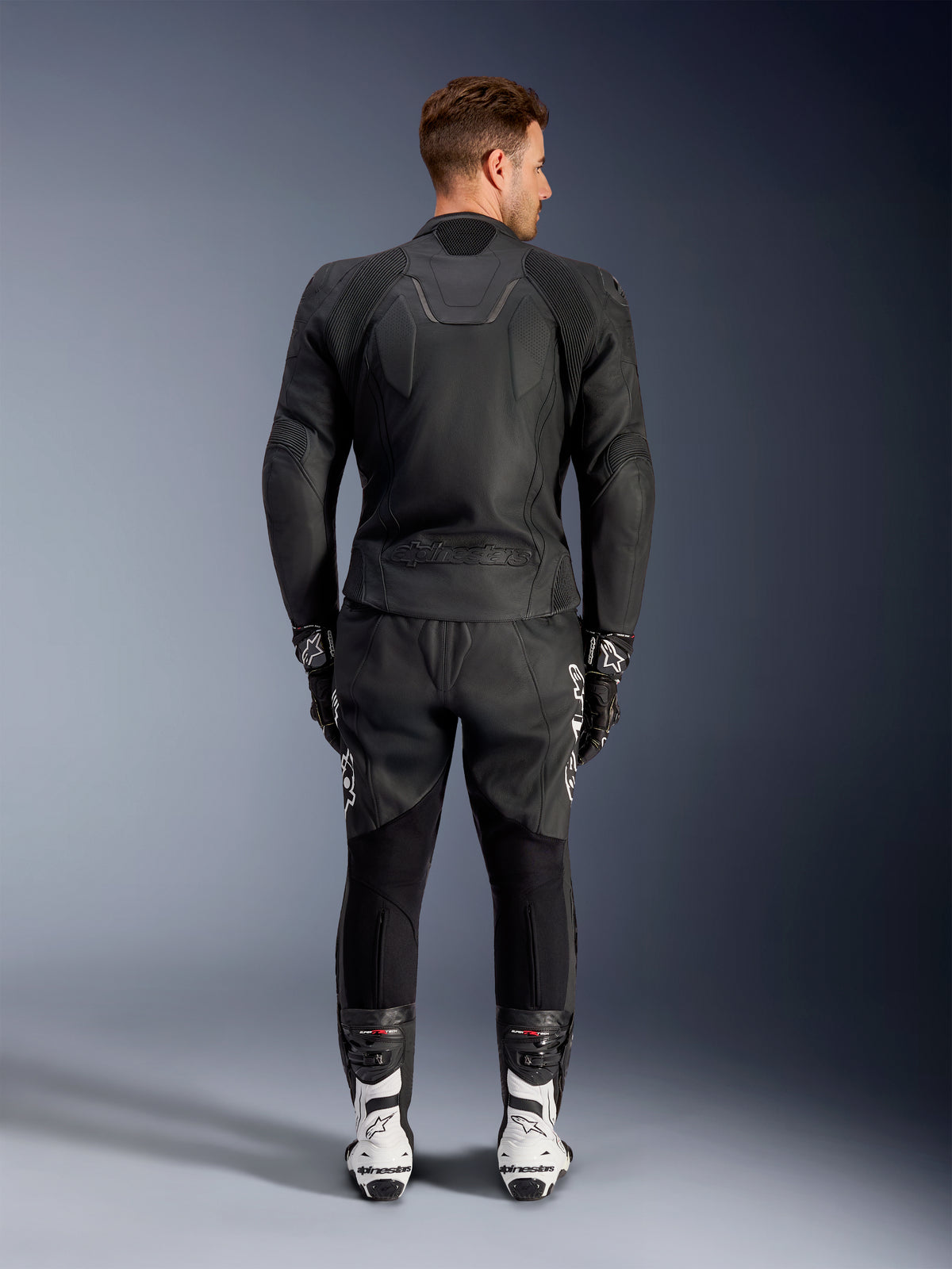 Vista posterior de un hombre que lleva la chaqueta de cuero Alpinestars Caliber en color negro, con un logotipo en relieve y paneles de cuero perforado, combinada con pantalones de cuero negros para motociclismo, guantes negros y botas de carreras blancas.