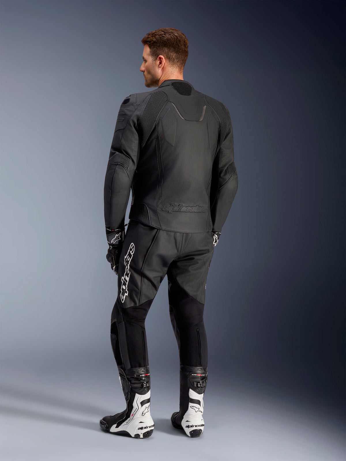 Chaqueta de cuero Alpinestars Caliber, chaqueta negra de moto para carretera, vista posterior, con cuero perforado e inserciones elásticas de acordeón en la cintura y los hombros, logotipo en relieve, usada con pantalones de cuero negros y botas de competición blancas