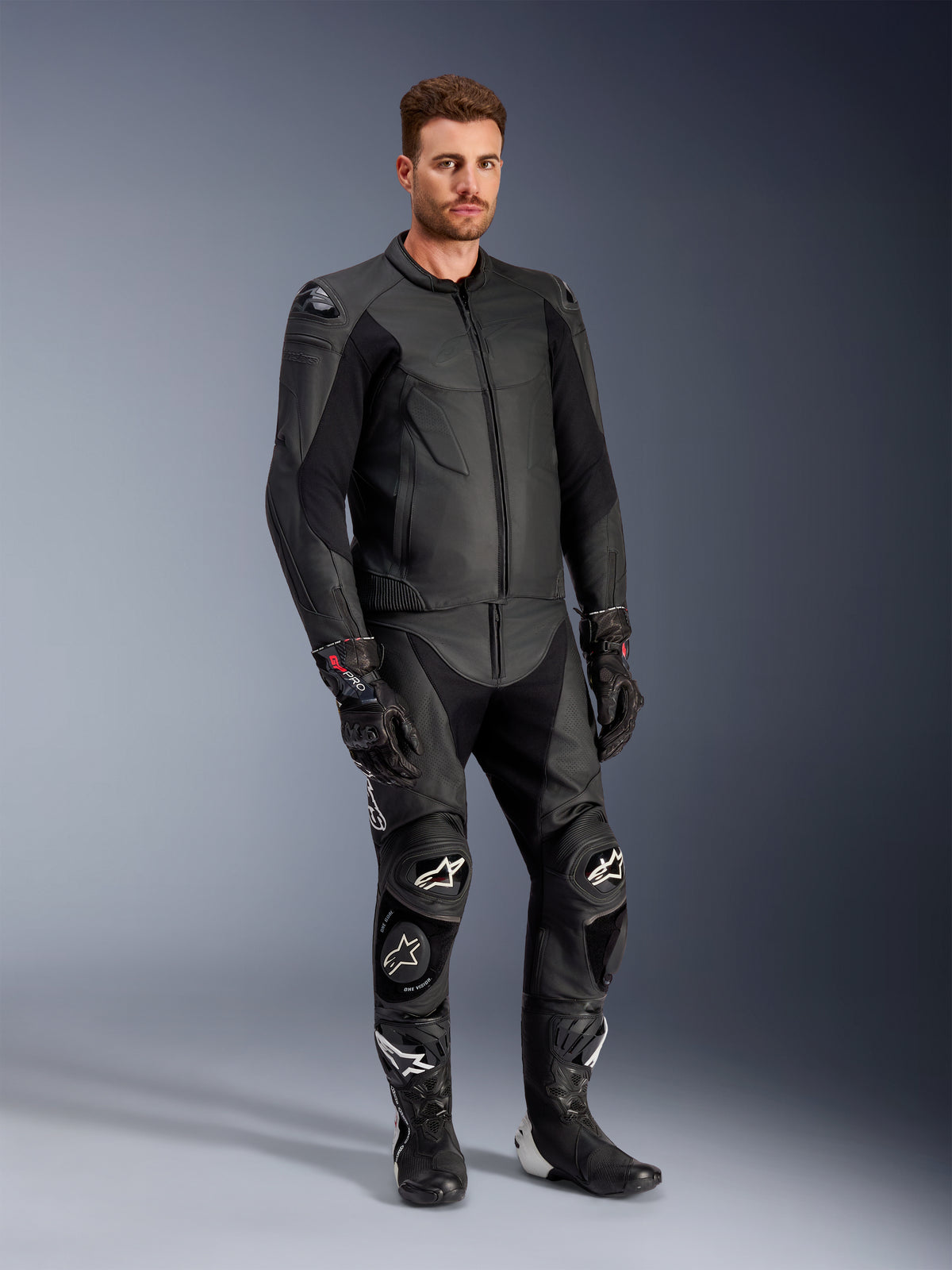 Modelo masculino luciendo la chaqueta de cuero Alpinestars Caliber en color negro, combinada con pantalones de moto de cuero negros a juego, guantes de competición y botas, mostrando un conjunto completo de equipamiento deportivo para carretera.