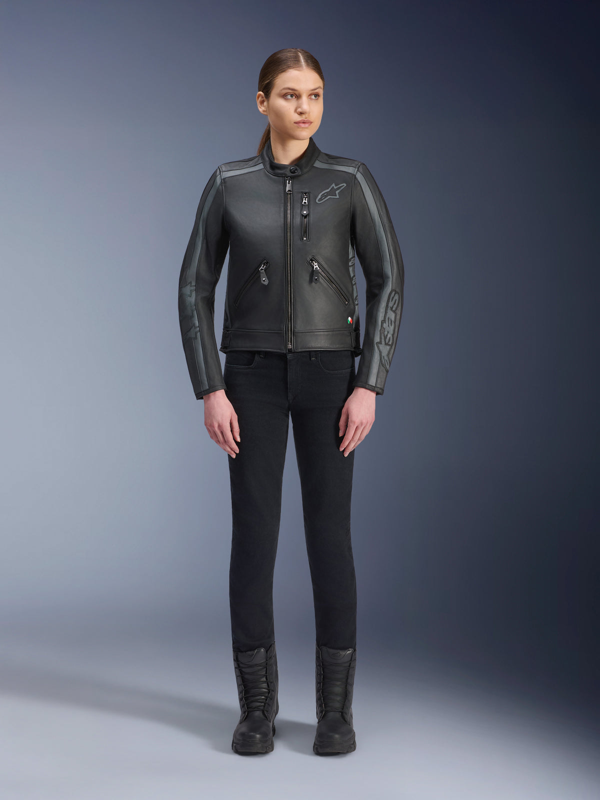 Chaqueta de cuero Alpinestars Stella Dyno, chaqueta de moto para mujer, color negro y gris oscuro, vista de estudio de cuerpo completo de una modelo usando la chaqueta con pantalones ajustados negros y botas negras, confeccionada en cuero de bovino con detalles grises y marca sutil de Alpinestars en el pecho y las mangas.