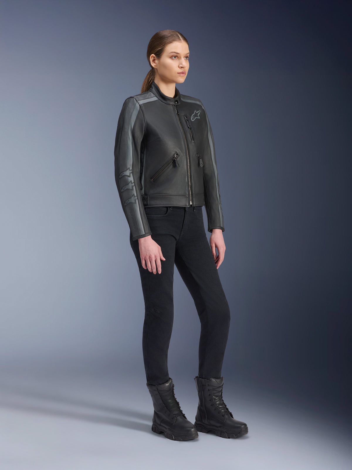 Chaqueta de cuero Alpinestars Stella Dyno, chaqueta de moto para mujer, color negro gris alquitrán y gris oscuro, combinada con pantalones de montar y botas negras, con rayas de carreras grises en las mangas y diseño de cuero tradicional
