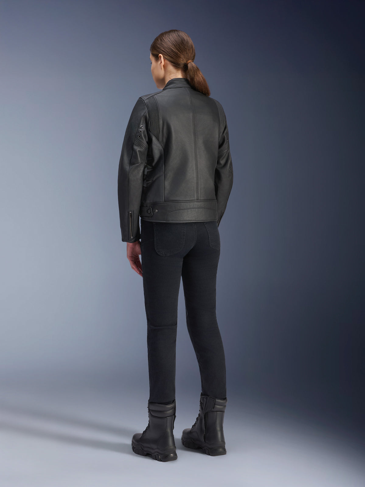 Chaqueta de cuero Alpinestars Stella Dyno, chaqueta de moto para mujer, negra, vista trasera que muestra la construcción de cuero de primera calidad y el ajuste entallado, usada con pantalones de montar negros y botas negras, estilo de conducción urbana.