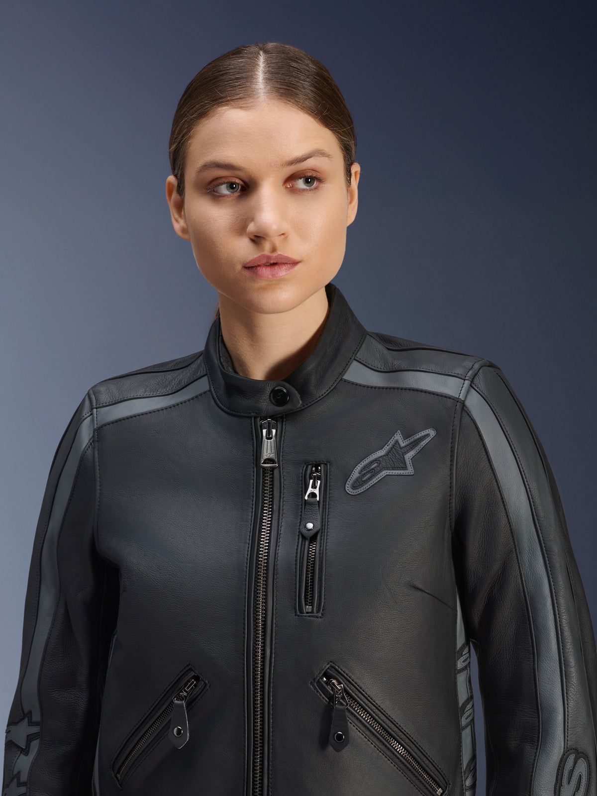 Modelo femenina luciendo la chaqueta de cuero Alpinestars Stella Dyno en Black Tar Gray Dark Gray, chaqueta de moto de cuero bovino de primera calidad con un diseño de herencia, con detalles grises en los hombros y múltiples bolsillos con cremallera, diseñada para un ajuste específico para mujer.