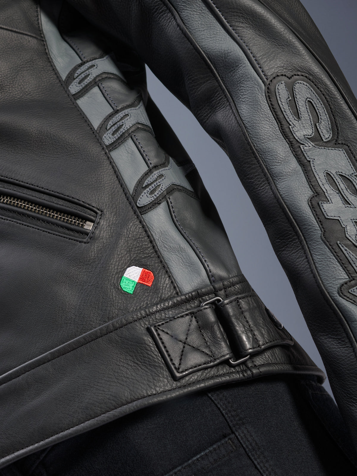 Alpinestars Stella Dyno Leather Jacket, detalle en cuero de bovino negro y gris Tar Gray, primer plano con bordado de la bandera italiana, hebilla de cintura ajustable y paneles de cuero gris con logos de Alpinestars y marca Stella en relieve en la manga.