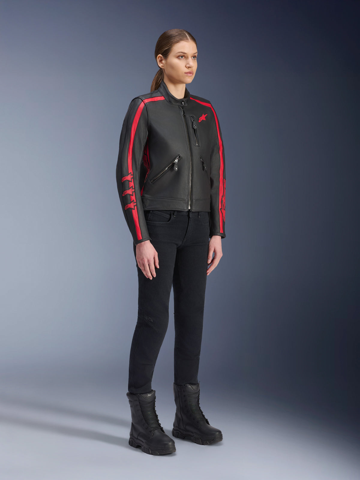 Modelo femenina con la chaqueta de cuero Alpinestars Stella Dyno en Black Haute Red, combinada con pantalones y botas negros, chaqueta de moto de cuero de estilo heritage con franjas rojas en los brazos y logotipo rojo, equipamiento de conducción urbana y de carretera para mujer