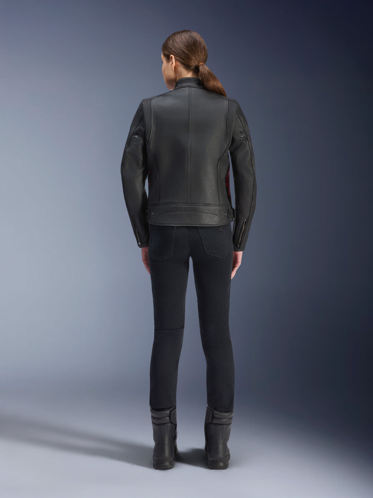 Vista posterior de una mujer luciendo la chaqueta de cuero Alpinestars Stella Dyno, negra con detalles en rojo Haute Red, combinada con pantalones negros ajustados y botas negras, diseño de chaqueta de cuero para moto de estilo heritage moderno