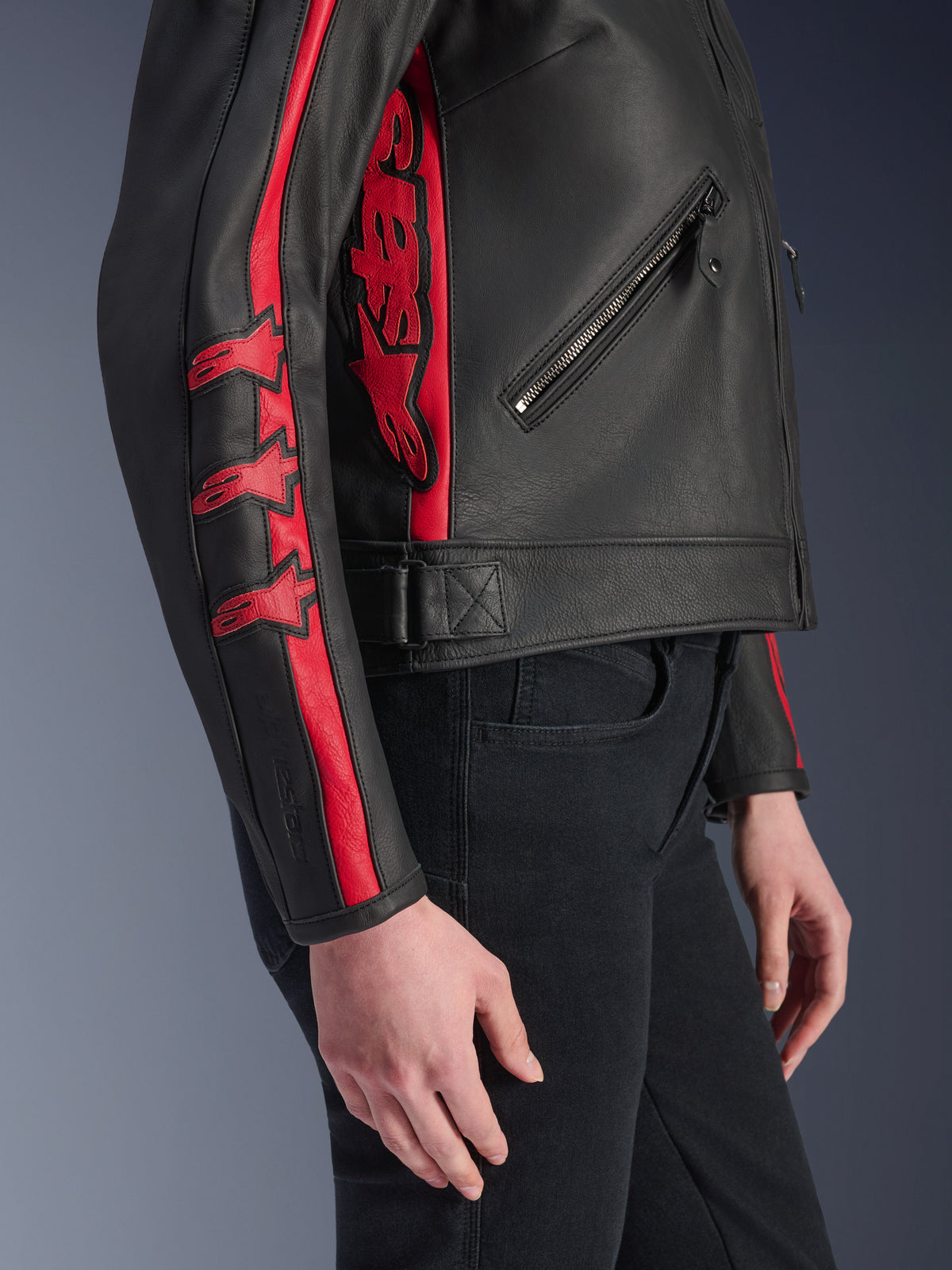 Chaqueta de cuero Alpinestars Stella Dyno, chaqueta de moto para mujer, negro y rojo Haute Red, vista lateral en una modelo, con franjas de carreras rojas y logotipos de estrellas en la manga, gran logotipo rojo en el lateral, cuero de bovino de primera calidad, estilo de conducción urbana