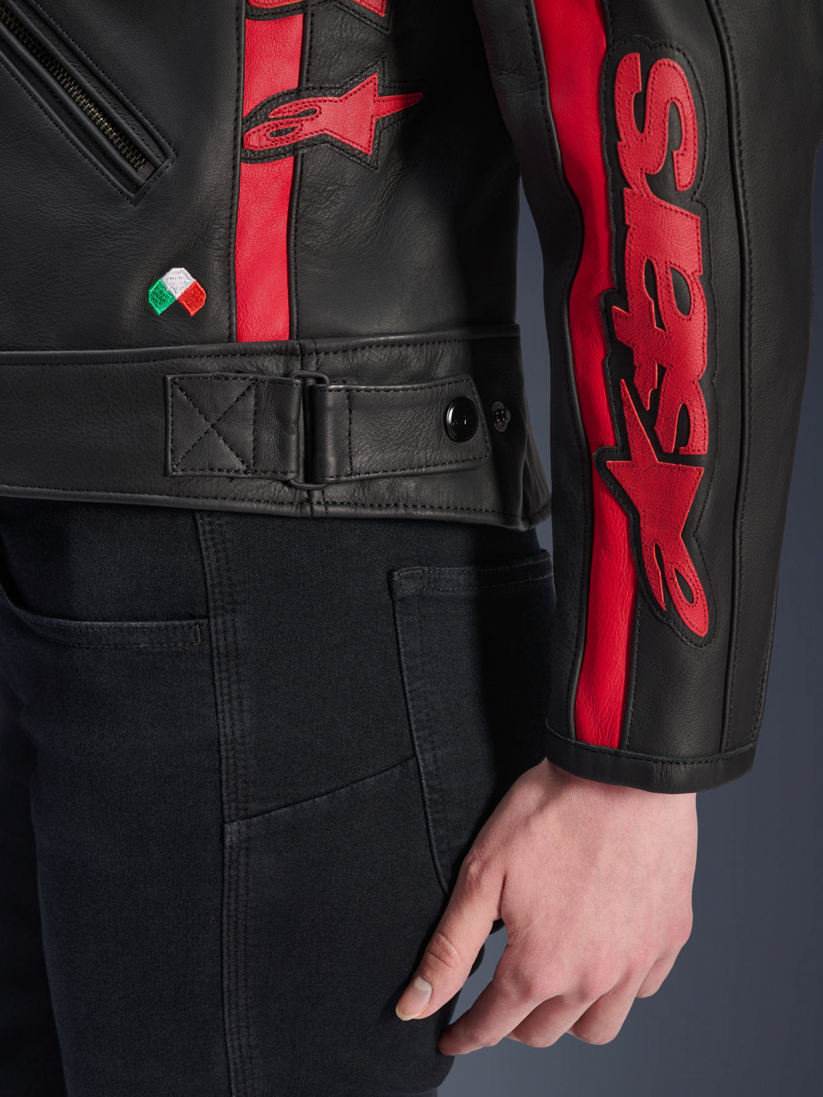 Chaqueta de cuero Stella Dyno de Alpinestars, chaqueta de moto de cuero para mujer, color negro y rojo, vista de cerca de la cintura y la manga, franjas rojas con el logotipo de Alpinestars, correa de ajuste en la cintura, pequeño bordado de la bandera italiana y confección en cuero bovino de primera calidad