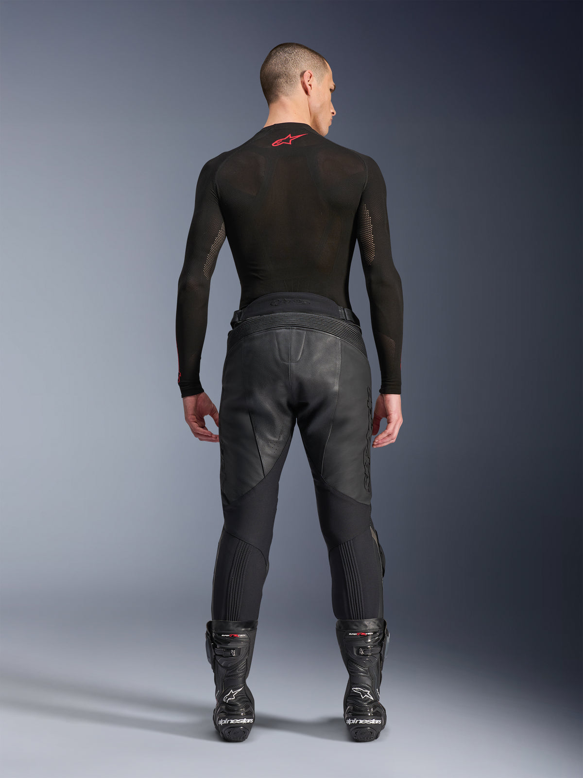 Alpinestars Missile V3 Leather Pants, negros, vista posterior en un modelo con camiseta de compresión de manga larga y botas de carreras, confección en cuero de primera calidad con superposición en la cintura alta y zonas elásticas, equipamiento profesional para carreras de motos
