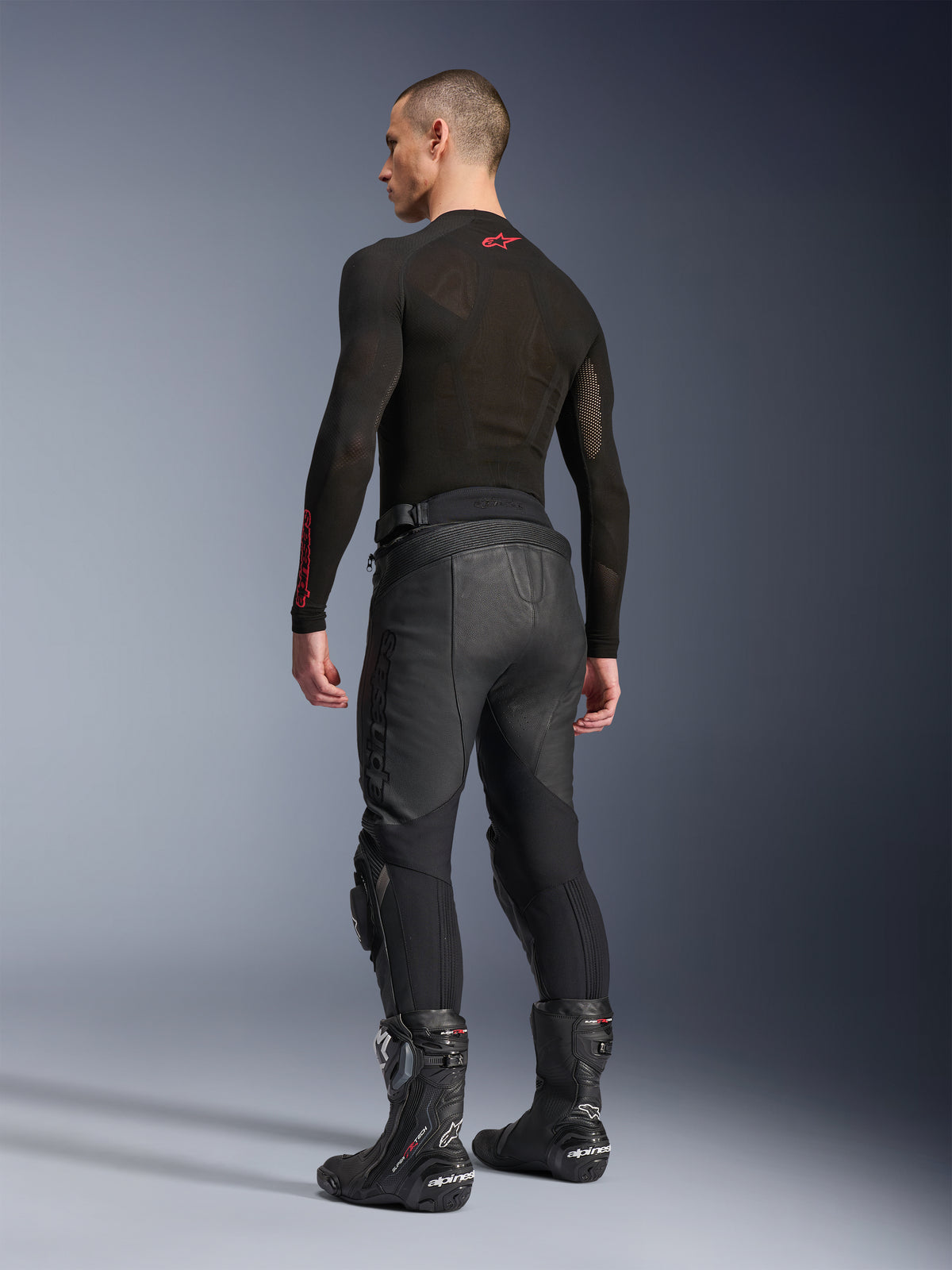 Pantalones de cuero Alpinestars Missile V3, negros, vista trasera en modelo, usados con una prenda base de manga larga negra y botas de carreras de moto negras, con paneles elásticos de cuero tipo acordeón y un diseño precurvado para un ajuste ergonómico al conducir.