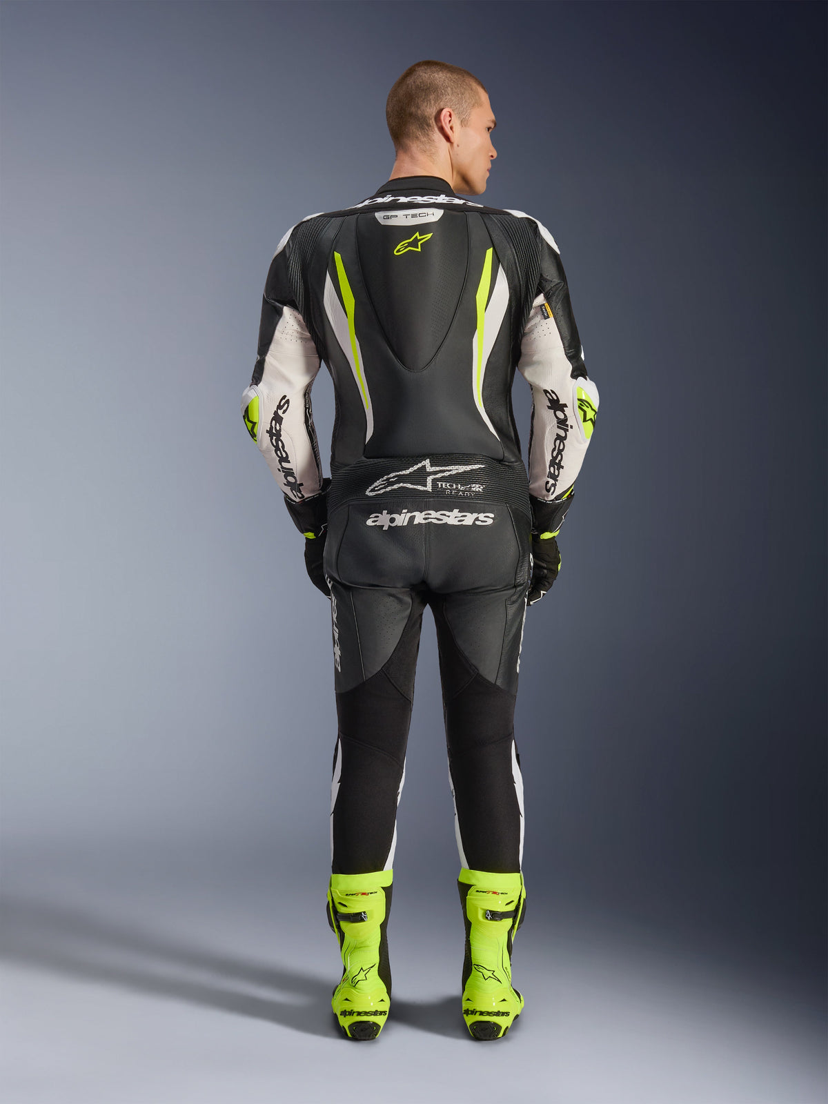 Mono de cuero de una pieza Alpinestars GP Tech V5, vista trasera, negro, blanco y amarillo flúor, con joroba aerodinámica con logo de estrella amarillo, marca Alpinestars en blanco en mangas y espalda, botas de competición en amarillo neón a juego, equipo profesional para circuito de motociclismo