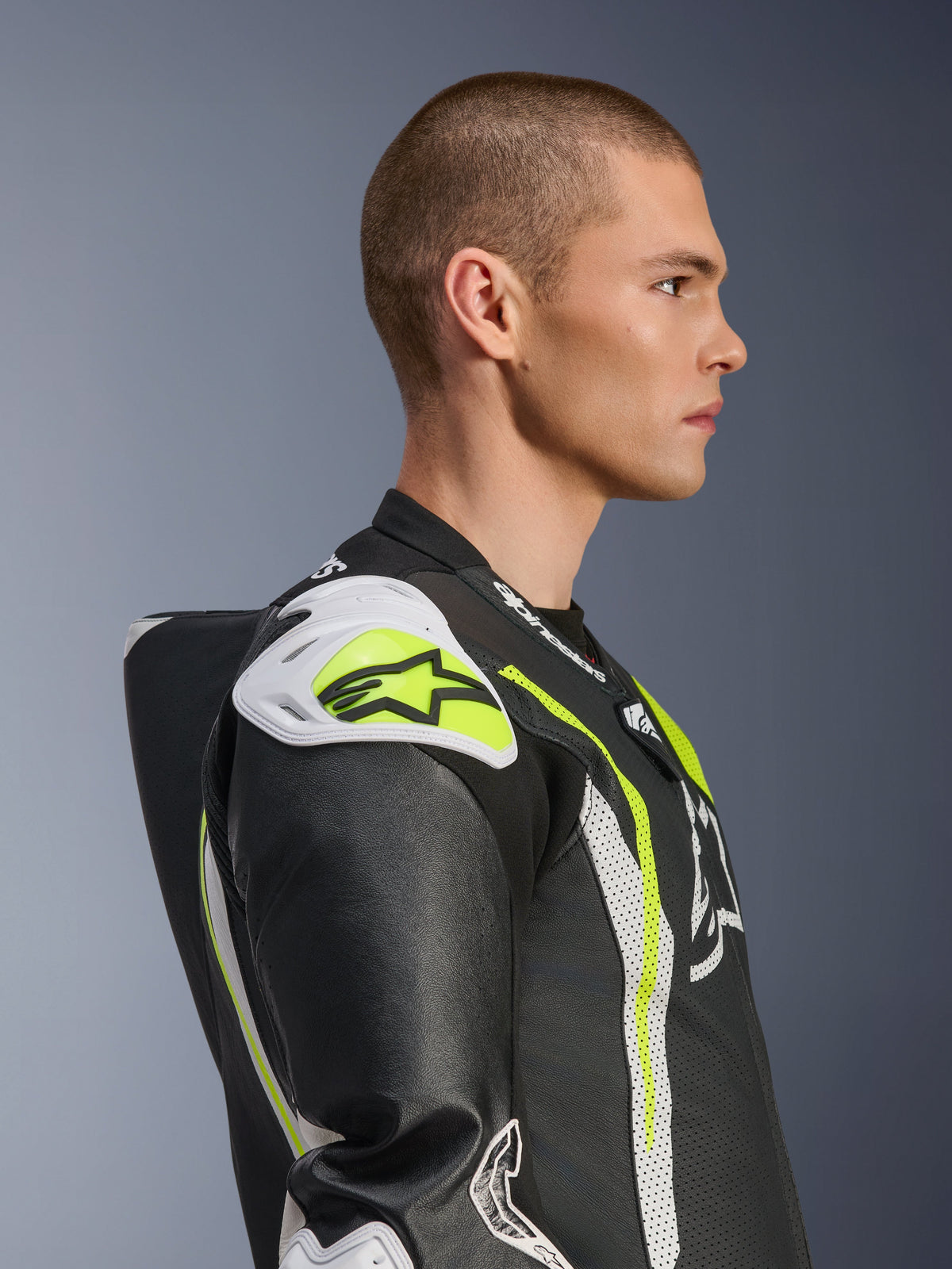 Modelo masculino con el mono de cuero de una pieza Alpinestars GP Tech V5 en negro, blanco y amarillo flúor, vista de perfil lateral que destaca la joroba aerodinámica y los deslizadores de hombro blancos, equipo de protección para moto inspirado en las carreras con detalles de cuero perforado