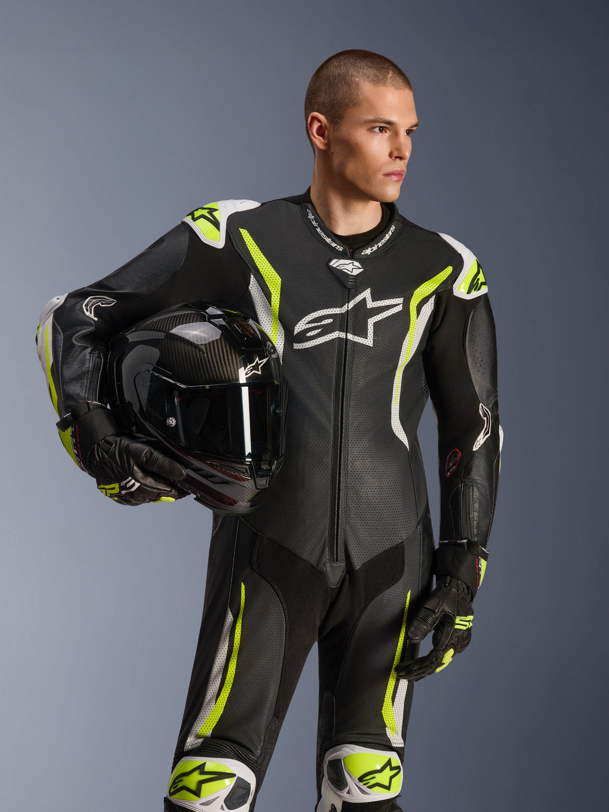 Mono de cuero de una pieza Alpinestars GP Tech V5, negro, blanco y amarillo flúor, modelo masculino sosteniendo un casco de moto negro, con paneles perforados, detalles en blanco y amarillo fluorescente y logotipo de Alpinestars destacado, equipo profesional de competición para motociclismo