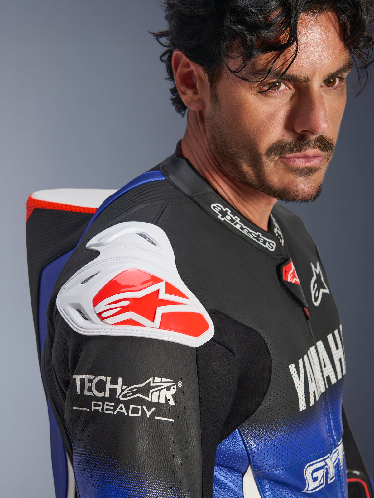 Yamaha GP-R7 One Piece Leather Suit, mono de cuero en azul metálico, negro, blanco y rojo, primer plano del hombro y el pecho, con protección de hombro DFS blanca con logo rojo, joroba aerodinámica y marca Tech-Air Ready en la manga de cuero perforado.