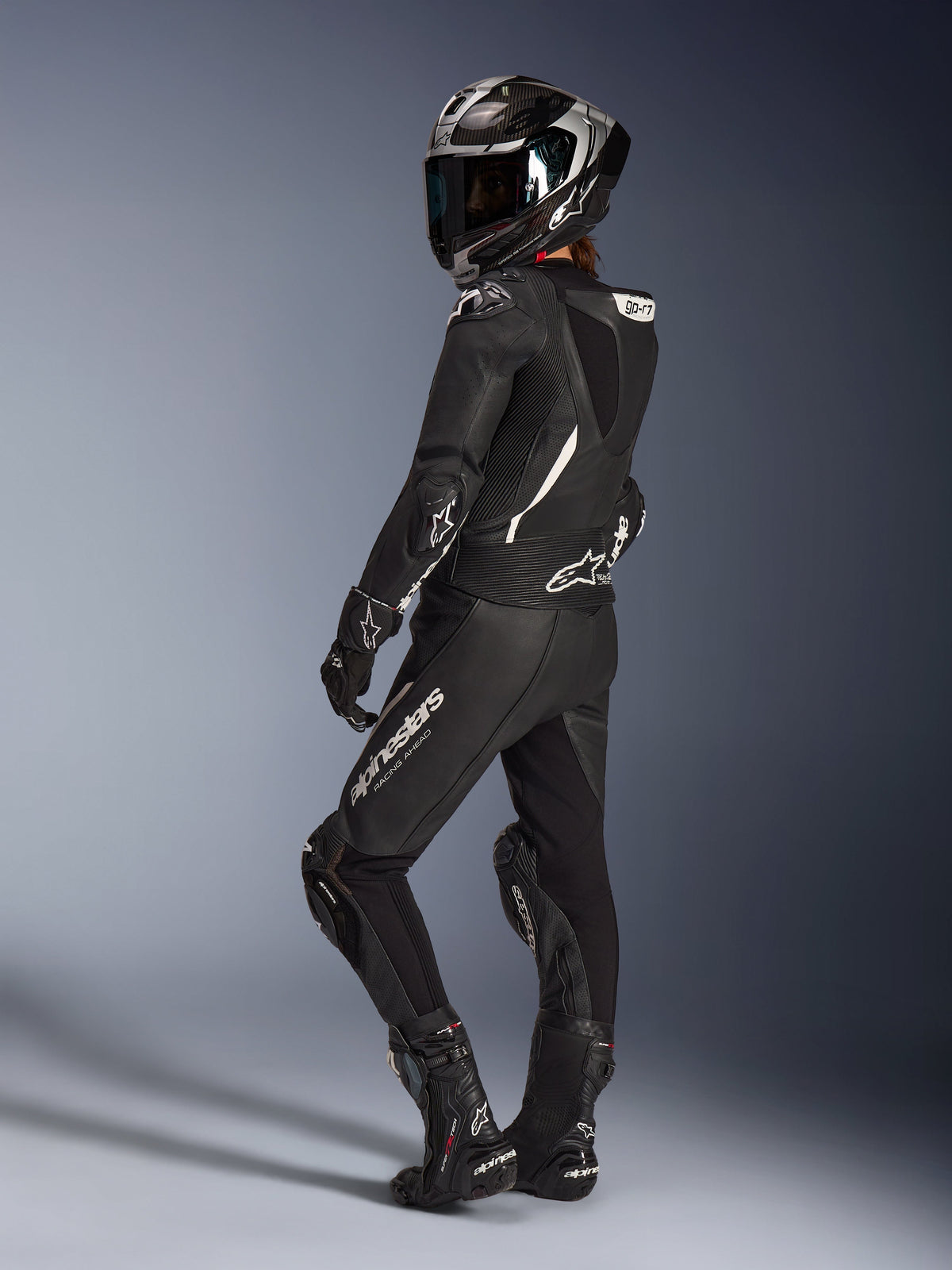 Piloto femenina con el traje de cuero de 2 piezas Alpinestars Stella GP-R7 en color negro, casco integral de carreras con acabado en carbono, guantes y botas a juego, vista trasera que muestra la joroba aerodinámica y los logotipos blancos de Alpinestars, equipamiento profesional para pista y conducción deportiva