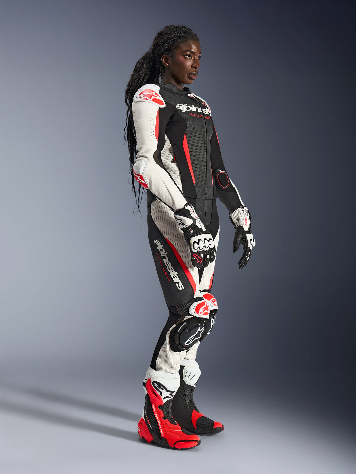 Piloto femenina vistiendo el traje de cuero de 2 piezas Alpinestars Stella GP-R7 en negro, blanco y rojo fluo, combinado con guantes y botas de carreras a juego, con paneles de cuero perforado y protecciones en hombros, codos y rodillas, de pie en un estudio.