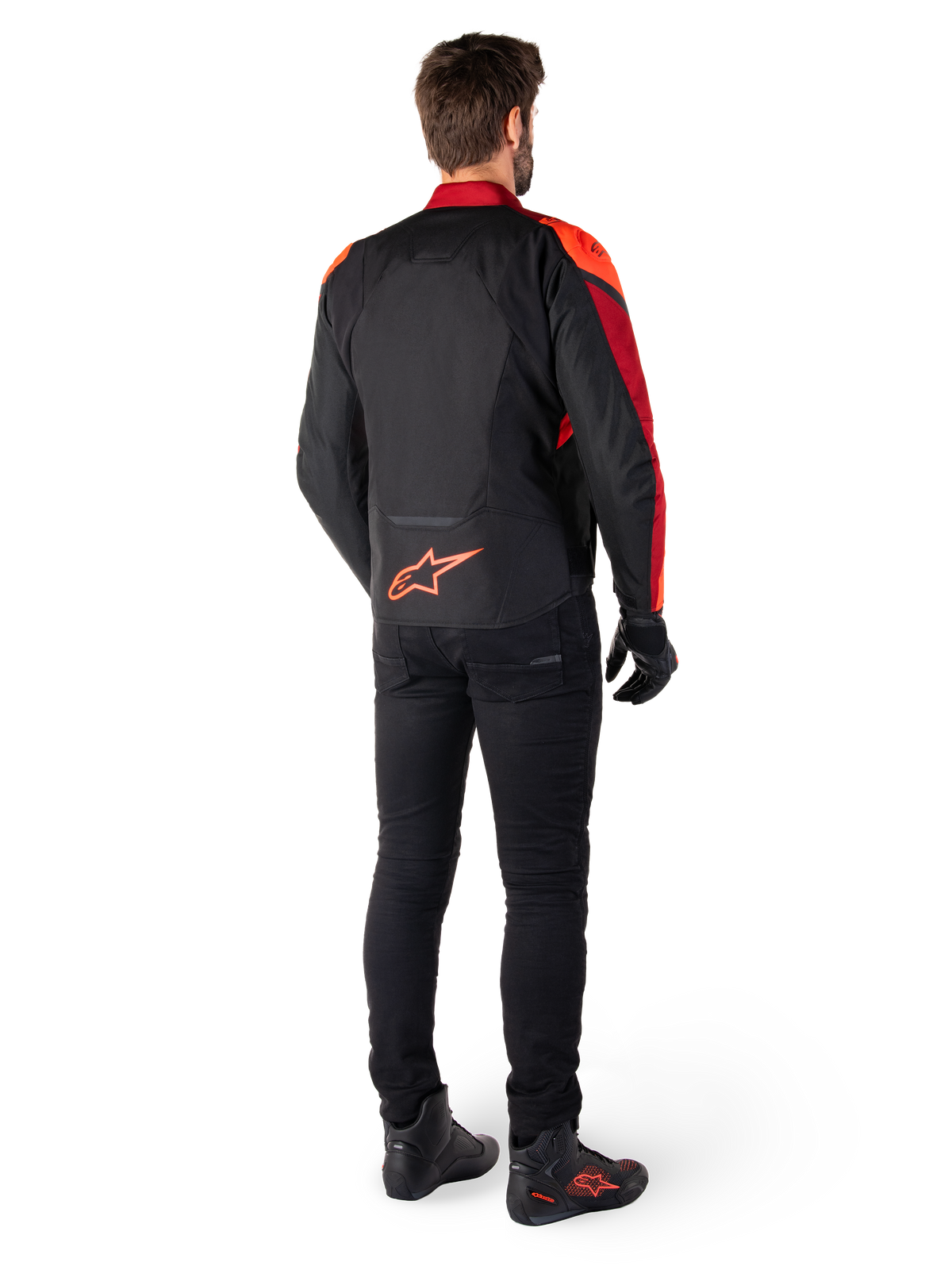 Chaqueta Alpinestars T-Jaws V4 Waterproof en color negro, rojo flúor y rojo oscuro, vista trasera de un piloto con la chaqueta textil, pantalones negros y botas de moto, con un llamativo logotipo rojo de Alpinestars en la parte inferior de la espalda.