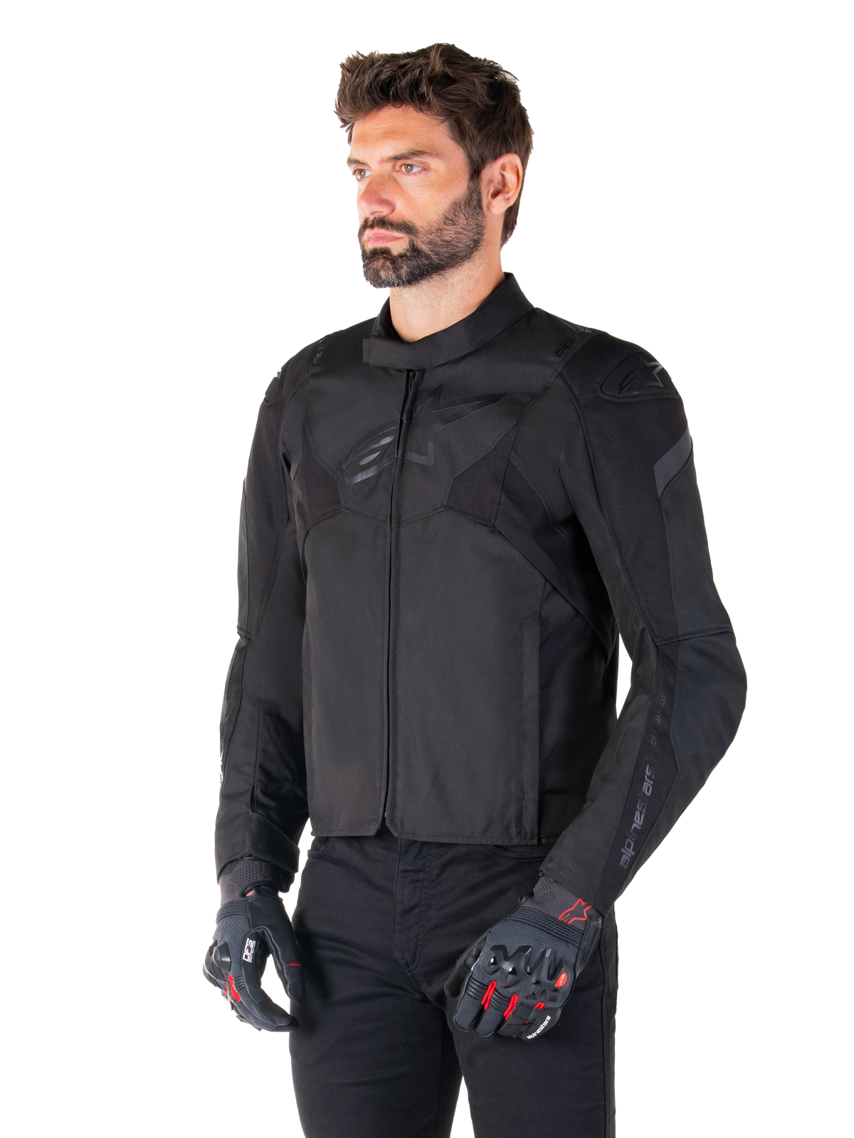 Piloto vistiendo la chaqueta impermeable Alpinestars T-Jaws V4 en color negro, combinada con pantalones negros y guantes protectores, con un ajuste deportivo estilizado, logotipos tonales de Alpinestars en el pecho y los hombros, y una confección textil de alto rendimiento.