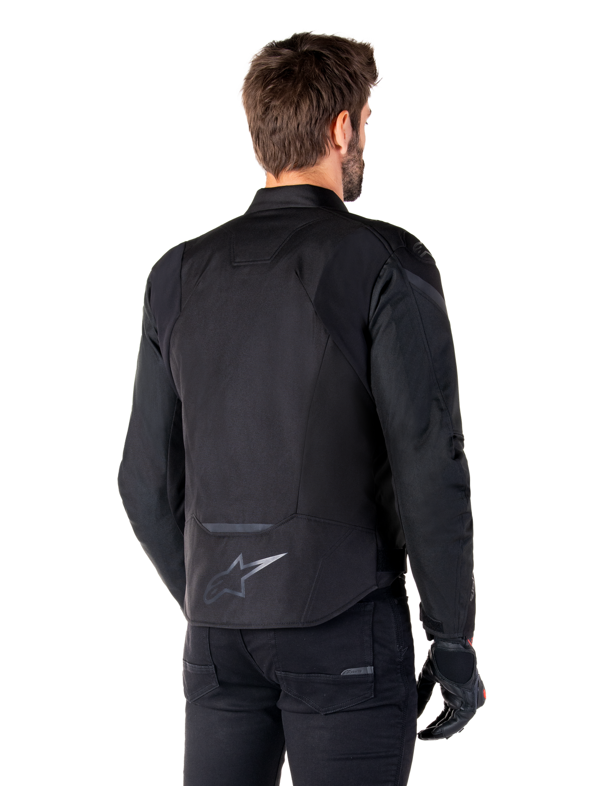 Chaqueta impermeable Alpinestars T-Jaws V4, negra, vista posterior en modelo, con logotipo reflectante de Alpinestars en la parte inferior de la espalda, parte trasera extendida para mayor cobertura, diseño textil deportivo con paneles tonales, combinada con guantes de moto negros
