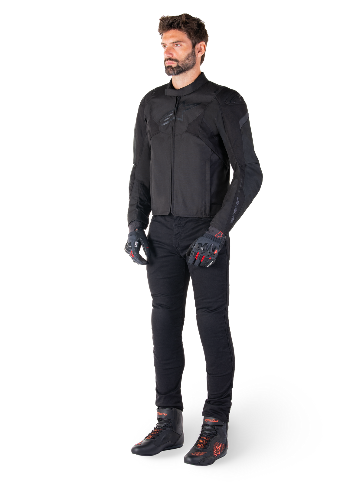 Modelo masculino vistiendo la chaqueta impermeable Alpinestars T-Jaws V4 en color negro, combinada con pantalones de conducción negros, guantes negros con detalles rojos y calzado de moto negro, con un ajuste deportivo elegante y logotipos sutiles, aislado sobre un fondo blanco.