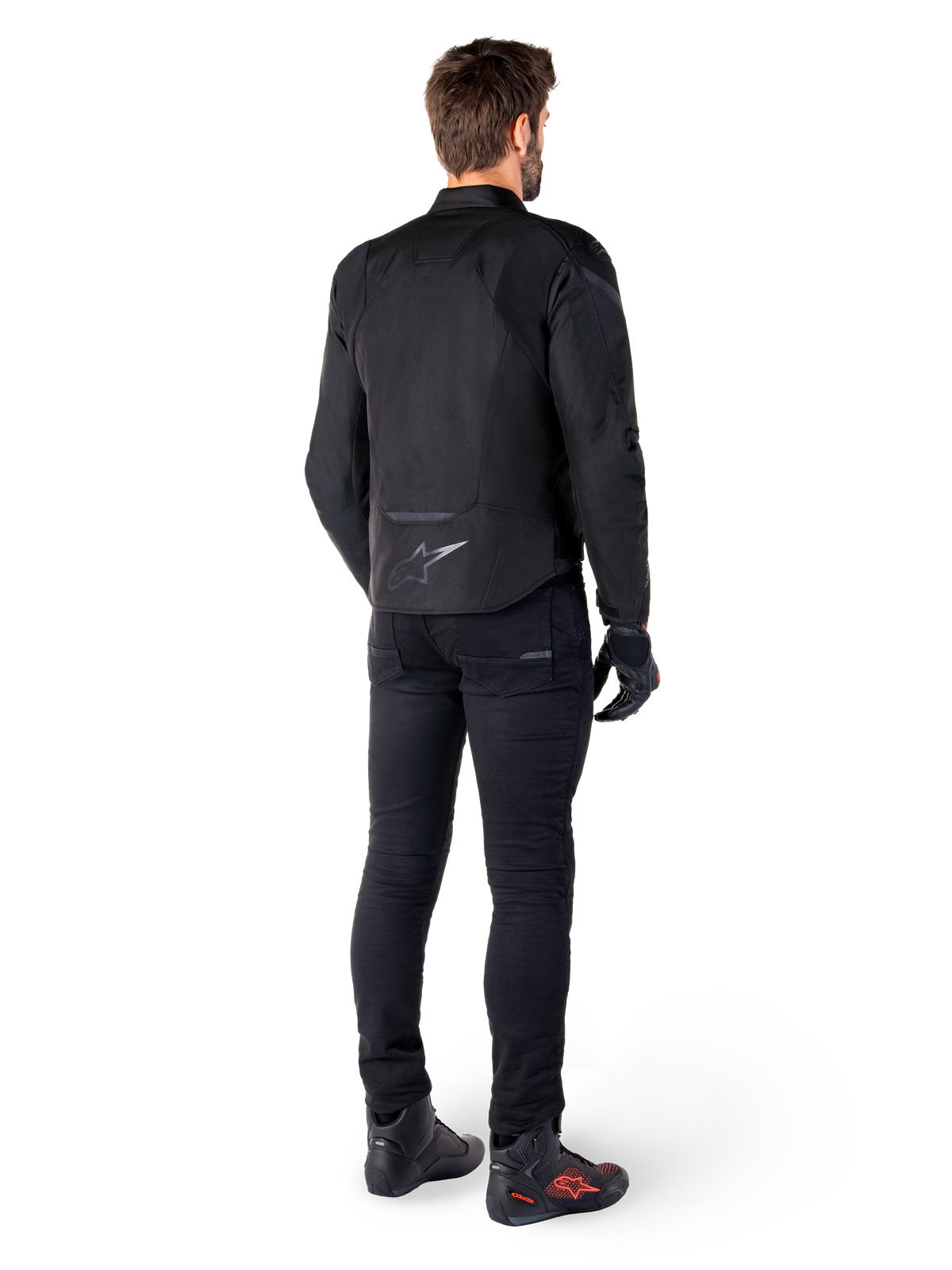 Chaqueta impermeable Alpinestars T-Jaws V4, negro, vista posterior en modelo con pantalones y botas de conducción negros, mostrando la construcción textil deportiva y el logotipo de Alpinestars en la parte inferior de la espalda.