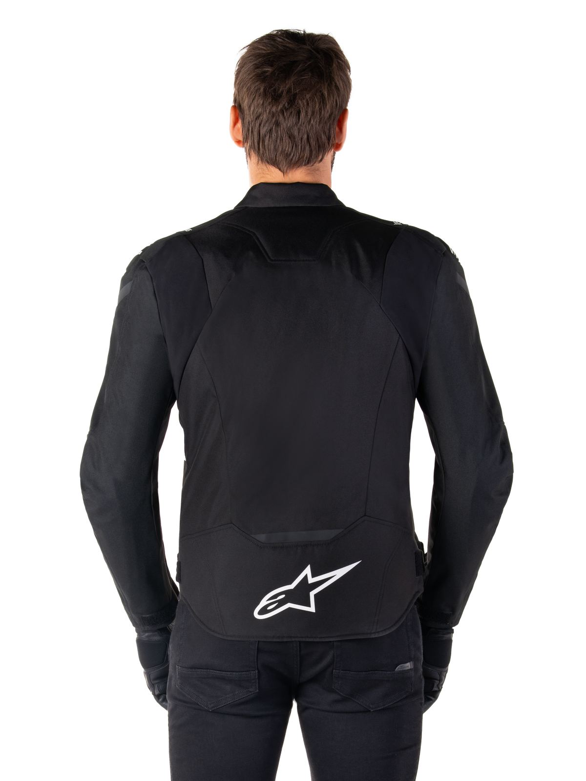 Chaqueta impermeable Alpinestars T-Jaws V4, chaqueta de moto negra vista desde atrás, con el logotipo blanco de Alpinestars en la parte inferior de la espalda, parte trasera extendida para mayor cobertura, usada por un modelo con pantalones y guantes negros, diseño textil deportivo y estilizado