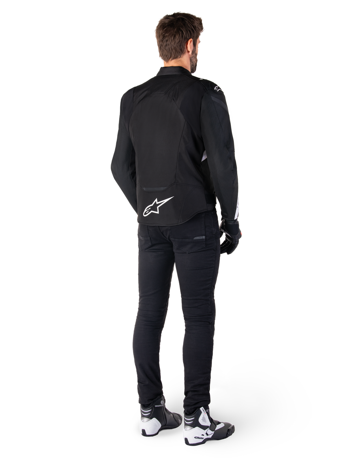 Chaqueta impermeable Alpinestars T-Jaws V4, negra y blanca, vista trasera que muestra el logotipo de la estrella blanca en la parte inferior de la espalda, usada por un modelo con pantalones de conducción negros, guantes negros y botas de moto blancas y negras.
