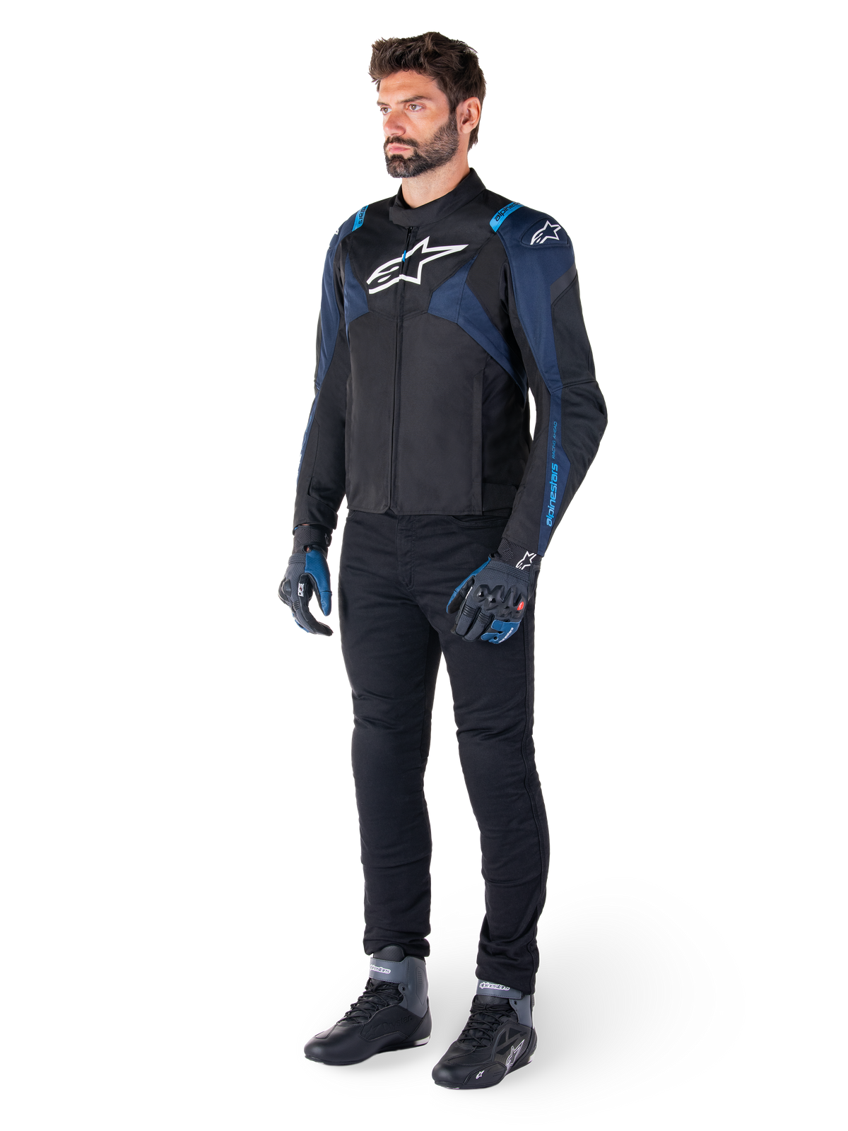 Chaqueta impermeable Alpinestars T-Jaws V4 en color negro, azul oscuro y azul Blithe, lucida por un modelo con pantalones de moto negros y botas de conducción, con logotipo blanco de Alpinestars en el pecho y detalles azules en los hombros, equipamiento de conducción de ajuste deportivo