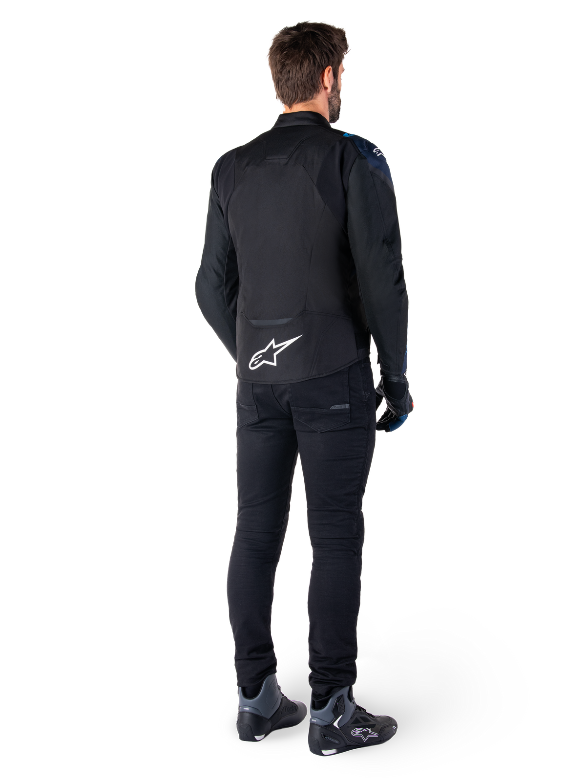 Vista posterior de un hombre con la chaqueta impermeable Alpinestars T-Jaws V4 en negro, azul oscuro y azul Blithe, con un destacado logotipo Astar blanco en la parte inferior de la espalda, combinada con pantalones negros y calzado para moto.