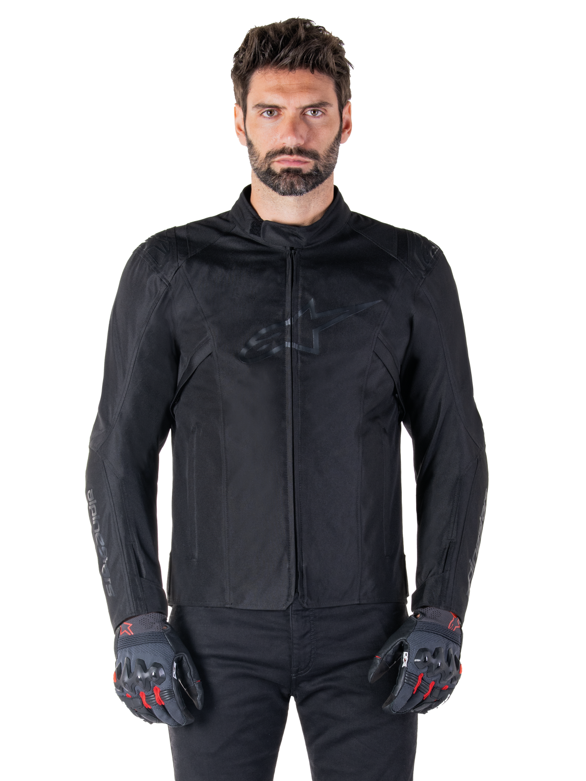 Alpinestars T-SPS V2 Waterproof Jacket, chaqueta de moto textil negra, usada por un piloto con guantes negros y rojos, con cuello alto, frontal con cremallera y un sutil logotipo en el pecho, protección impermeable para conducción deportiva y urbana