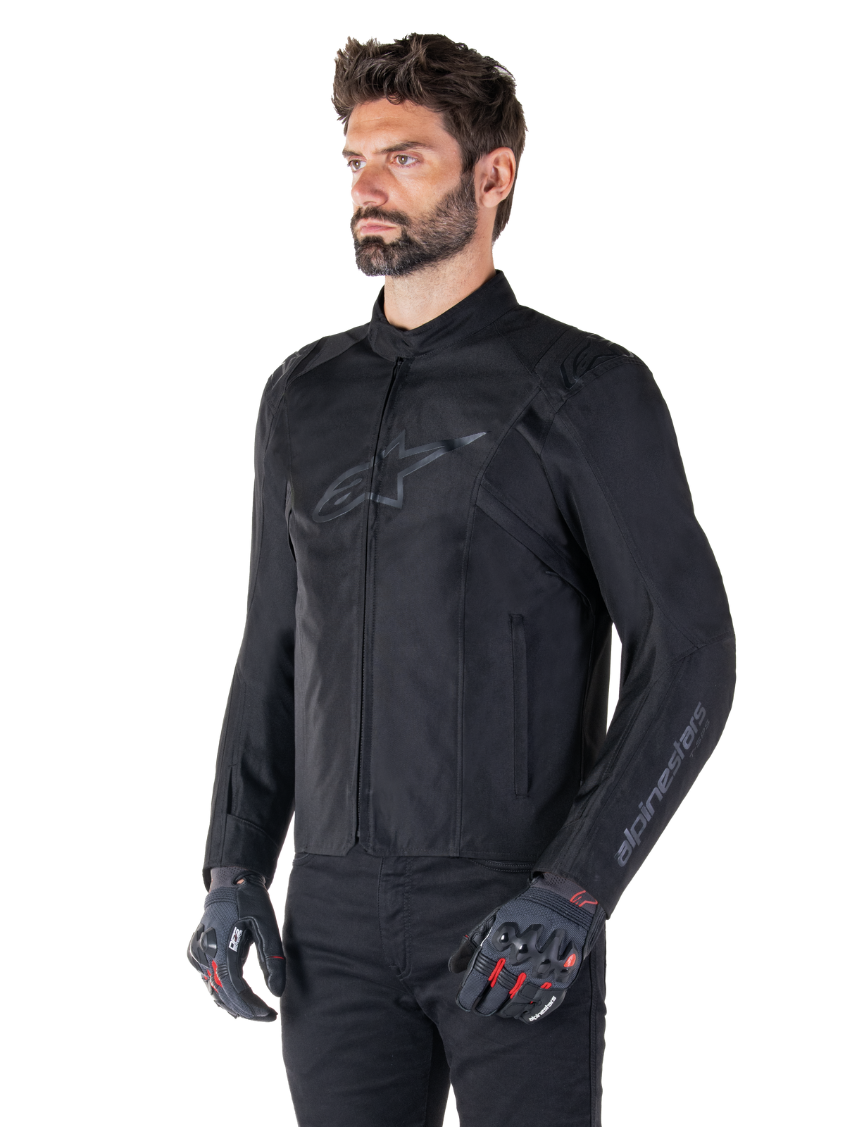 Modelo con la chaqueta impermeable Alpinestars T-SPS V2 en color negro, combinada con pantalones negros y guantes de moto grises con detalles en rojo, con un sutil logotipo negro en el pecho y marca en la manga, chaqueta textil de corte deportivo para conducción en carretera.