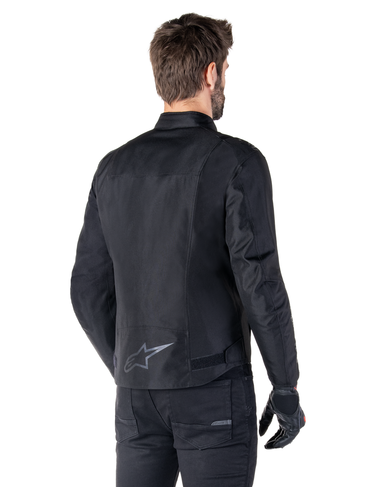 Chaqueta impermeable Alpinestars T-SPS V2, negro, vista posterior en un modelo masculino, con logotipo de estrella reflectante en la parte inferior de la espalda, perfil trasero extendido y correas de ajuste en la cintura, combinada con pantalones y guantes de conducción negros
