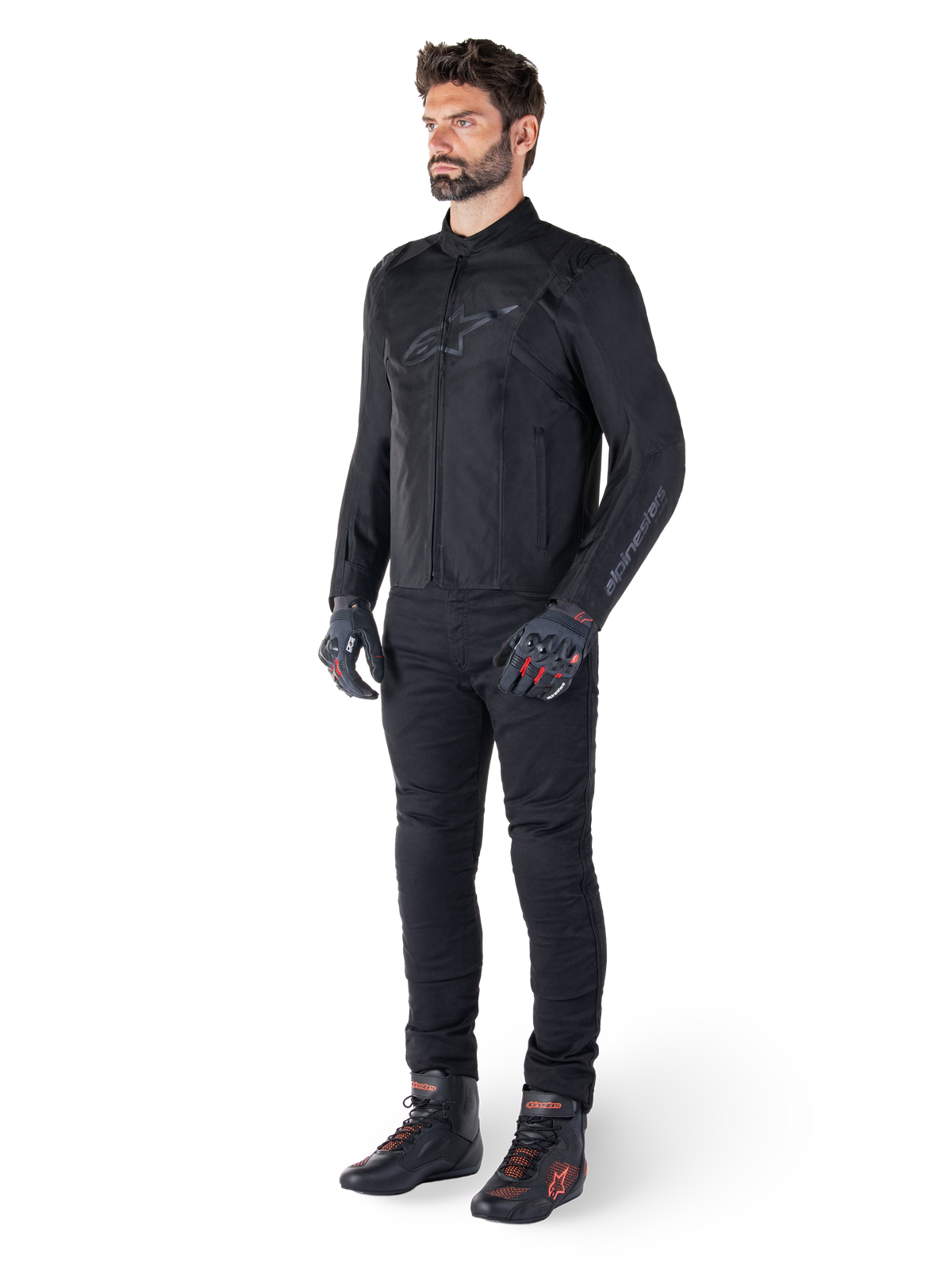 Chaqueta impermeable Alpinestars T-SPS V2, negra, usada por un modelo masculino con pantalones negros a juego, guantes negros con detalles rojos y calzado de conducción negro, equipamiento de moto completo para conducción urbana y deportiva, fondo blanco