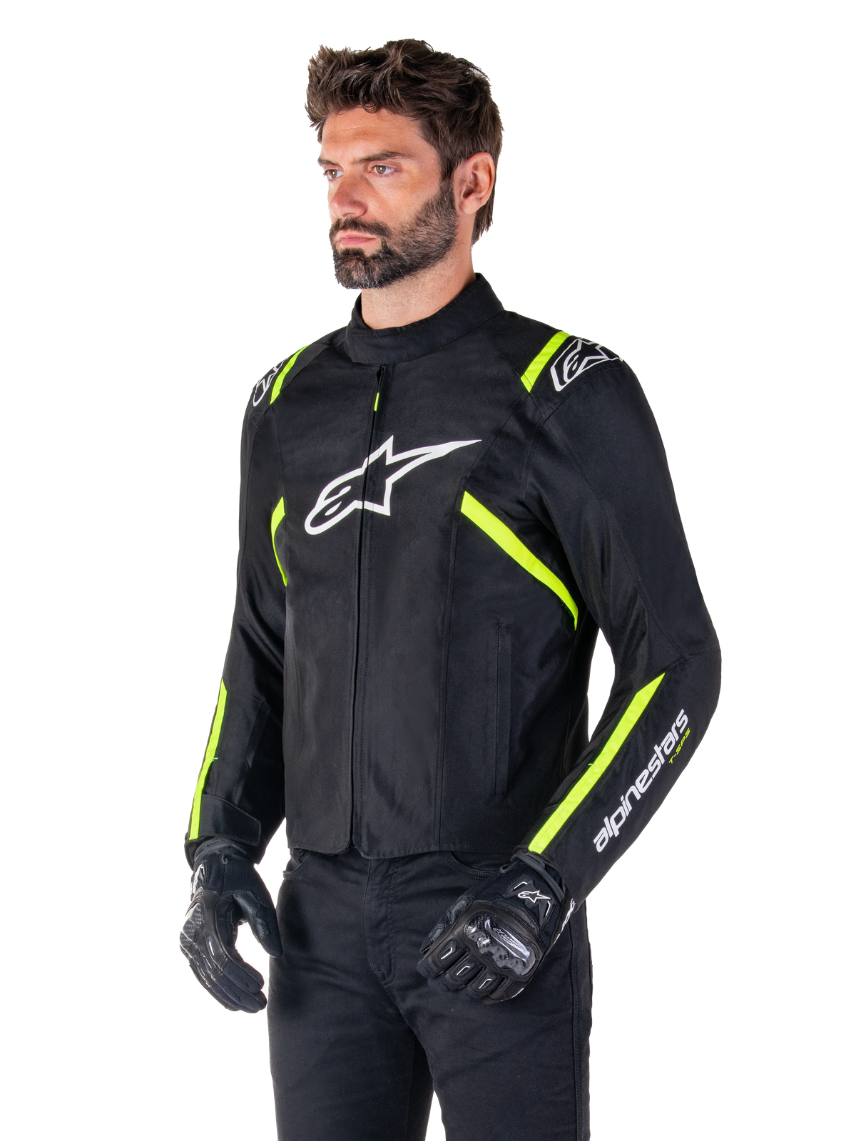 Modelo masculino con la chaqueta Alpinestars T-SPS V2 Waterproof, negra con detalles en blanco y amarillo flúor, con el logotipo Astars blanco en el pecho y franjas amarillo flúor en hombros y mangas, combinada con pantalones negros y guantes de conducción, chaqueta textil de moto de corte deportivo.