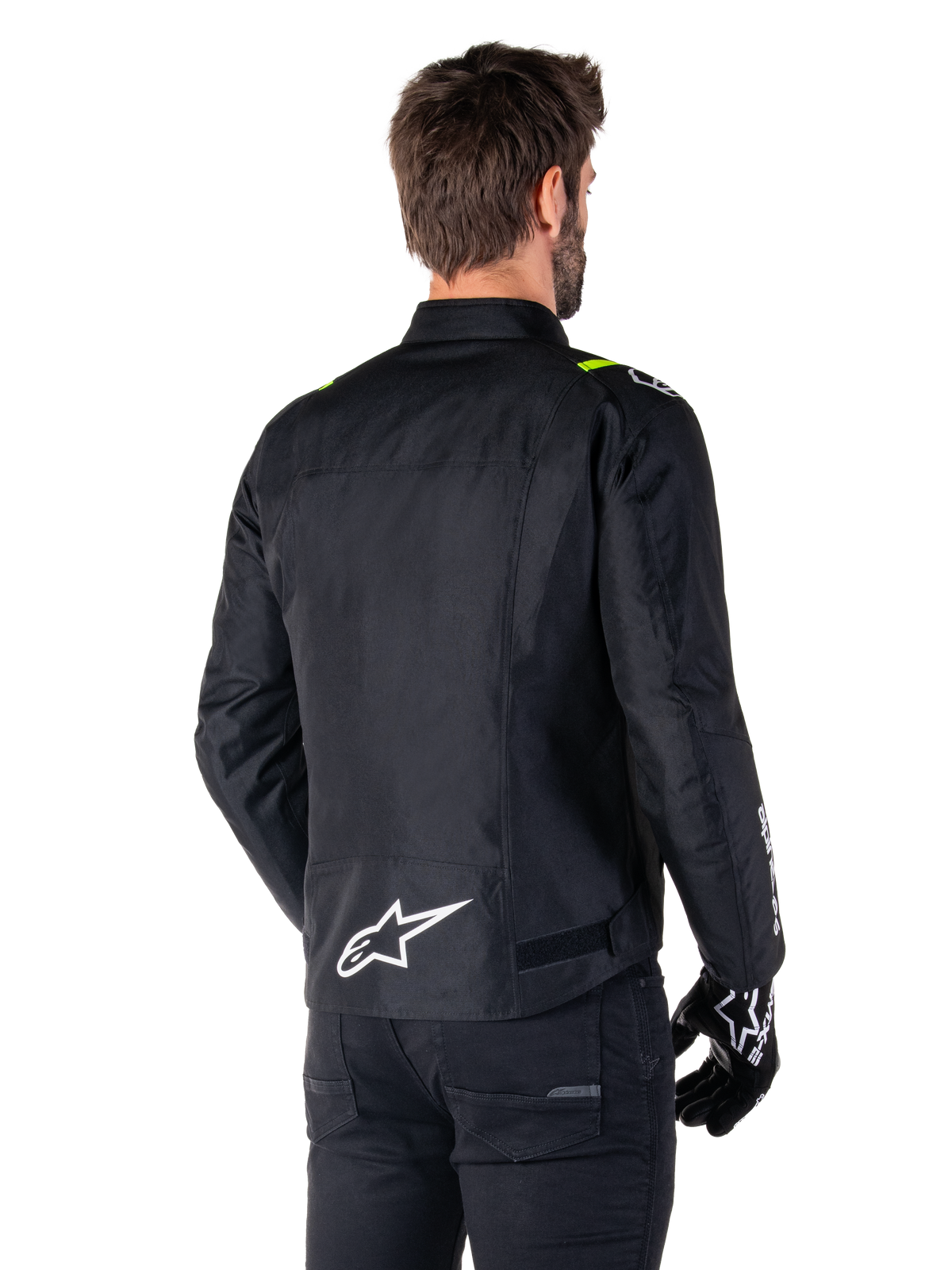 Chaqueta impermeable Alpinestars T-SPS V2, Negro Blanco Amarillo Flúor, vista trasera de modelo masculino vistiendo la chaqueta textil de moto, con un logotipo blanco en la parte inferior de la espalda y detalles en amarillo flúor en los hombros, combinada con pantalones y guantes negros.