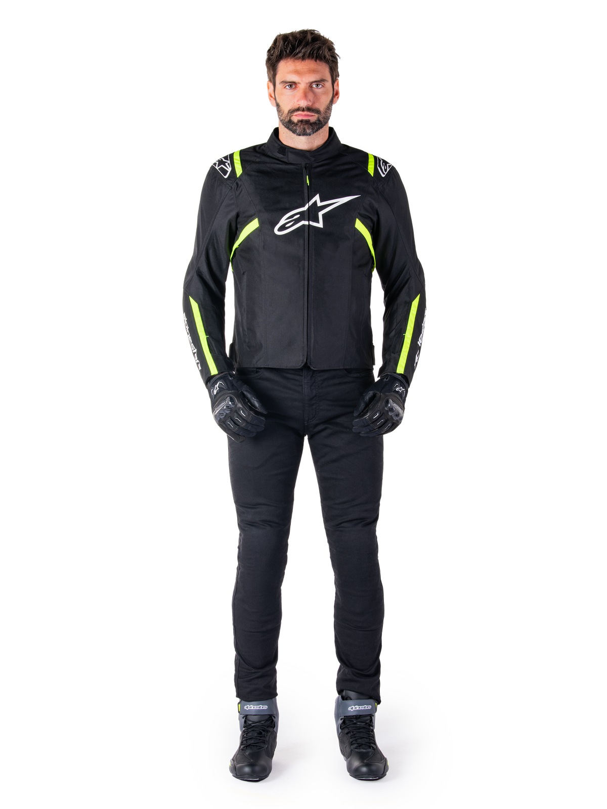 Chaqueta impermeable Alpinestars T-SPS V2, negro, blanco y amarillo flúor, lucida por un modelo con pantalones de moto y guantes negros, con logotipo de Alpinestars blanco en el pecho y detalles fluorescentes en hombros y mangas, equipamiento de motociclismo de corte deportivo
