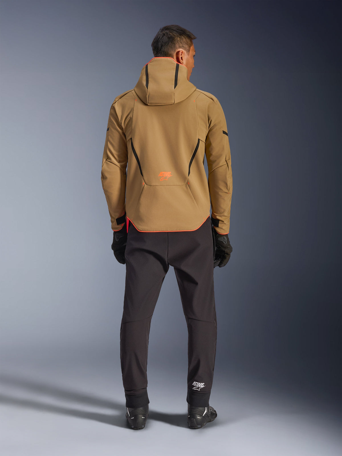 Chaqueta impermeable Alpinestars Aeroshell en Dark Khaki Black Orange Fluo, vista trasera de un hombre con la chaqueta softshell con capucha, con ribetes negros y logotipo naranja, combinada con pantalones técnicos negros y guantes