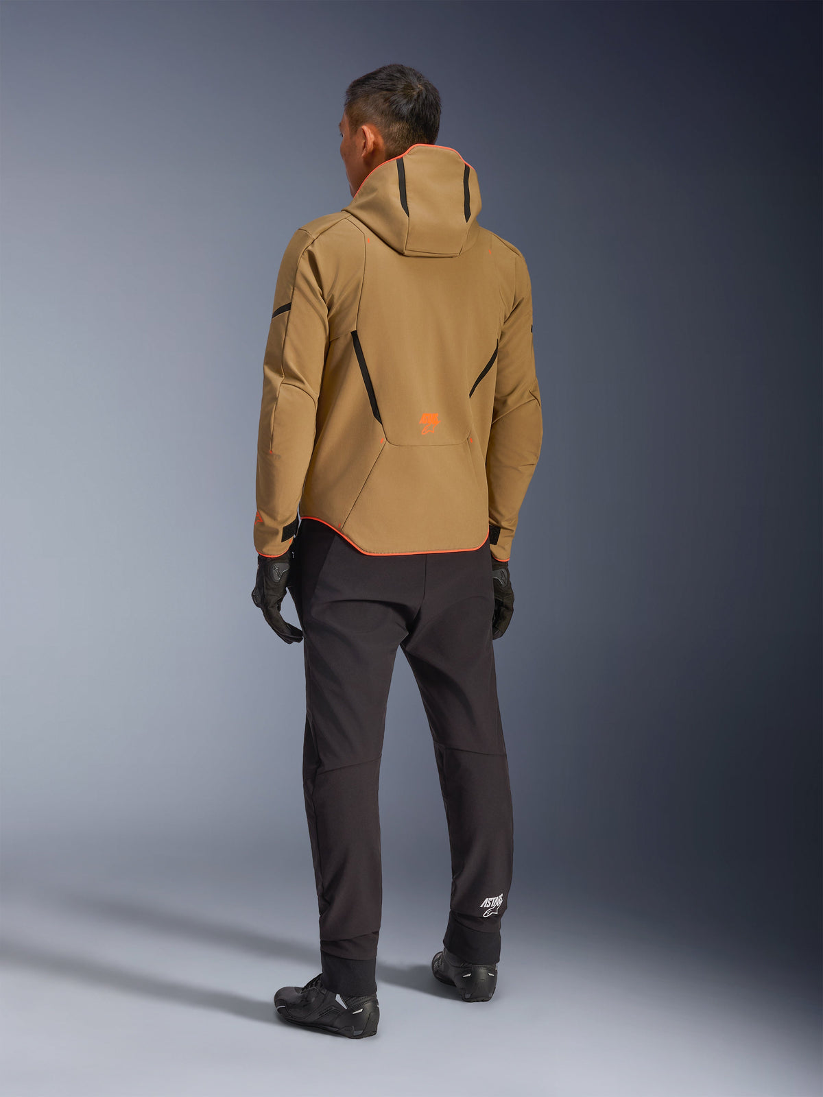 Chaqueta Alpinestars Aeroshell WP, caqui oscuro, negro y naranja flúor, vista posterior de un piloto con la chaqueta softshell con capucha, combinada con pantalones y guantes negros, con detalles geométricos negros y un logotipo naranja en la parte inferior de la espalda.
