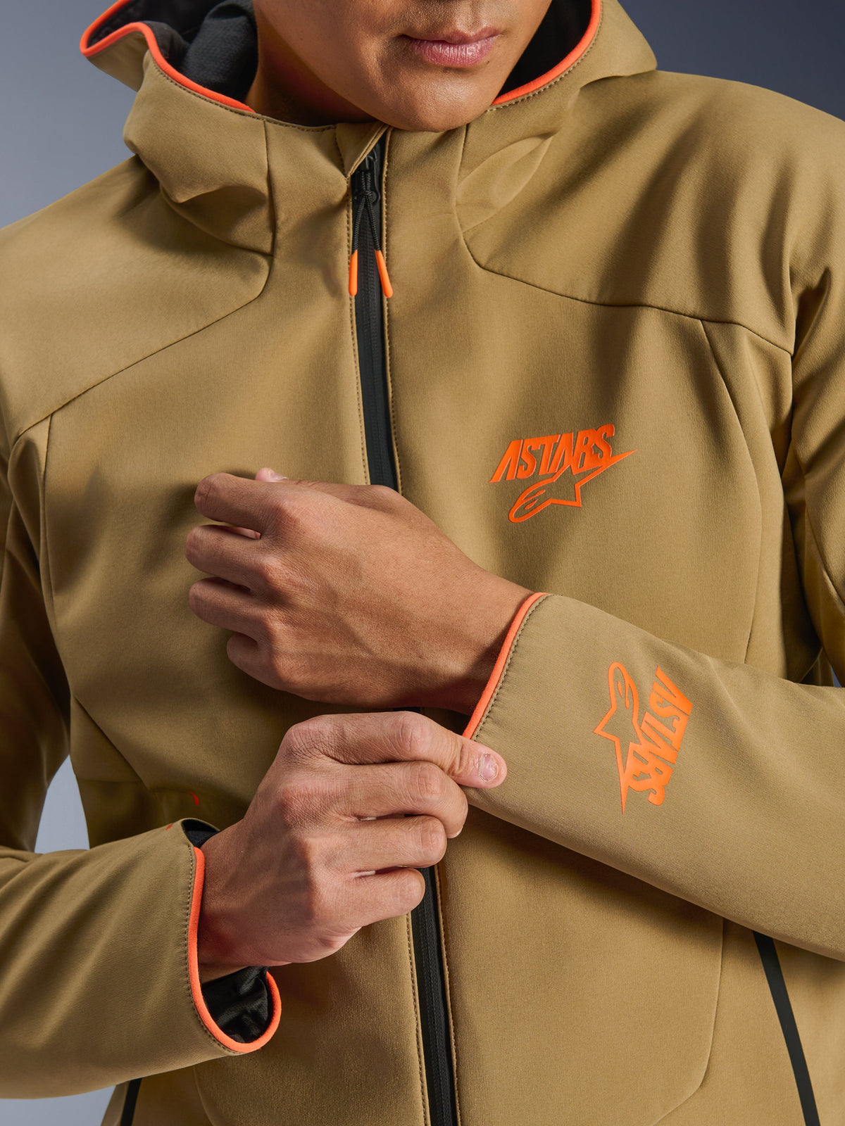 Chaqueta impermeable Alpinestars Aeroshell, caqui oscuro y naranja flúor, primer plano del frente y la manga con logotipos ASTARS en naranja, ribetes naranjas en la capucha y los puños, y una cremallera impermeable negra.
