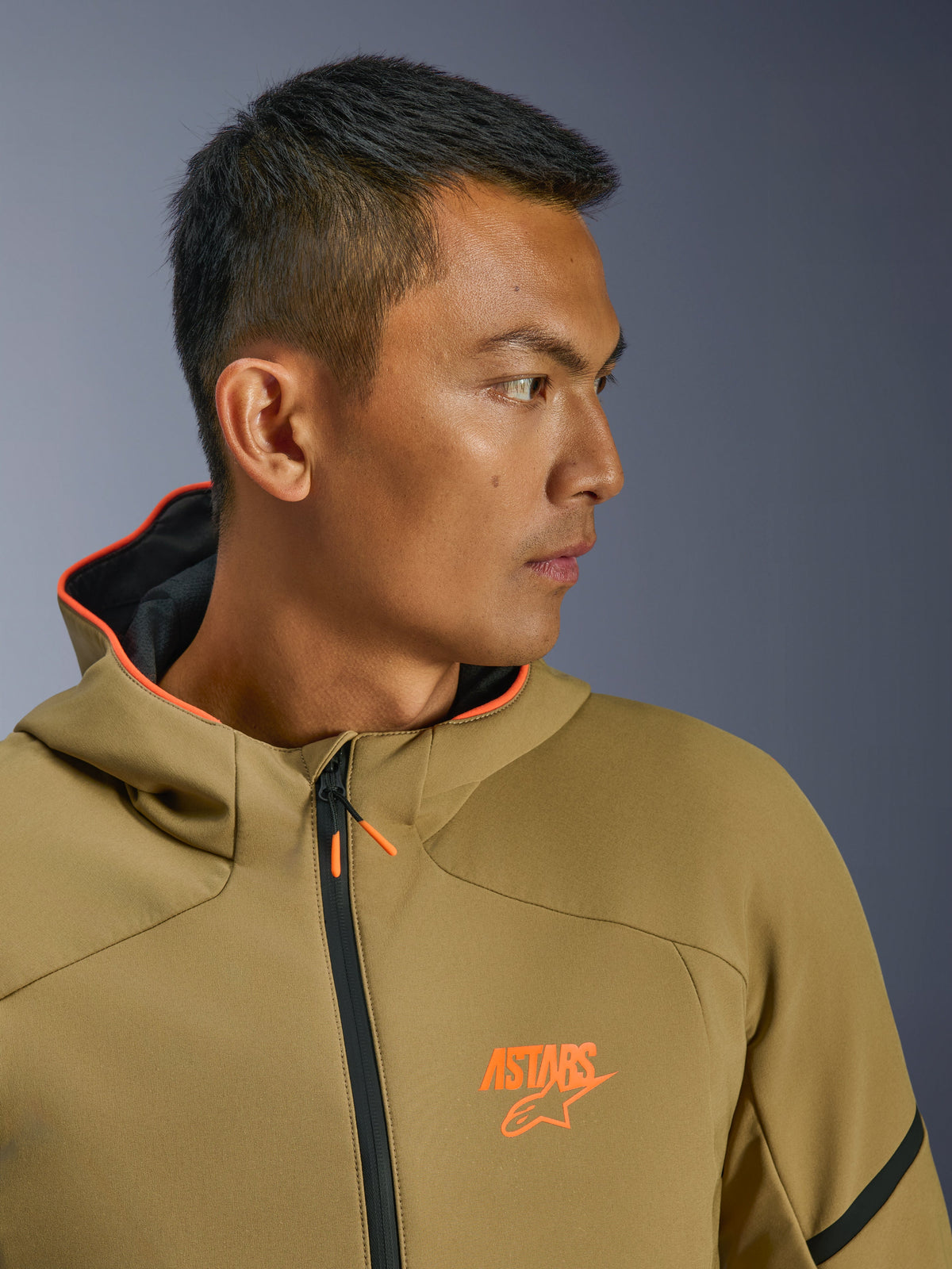 Chaqueta impermeable Alpinestars Aeroshell, caqui oscuro y naranja flúor, primer plano de perfil lateral, con logotipo ASTARS naranja en el pecho y capucha con ribete naranja, sudadera softshell moderna crossover para montar en moto