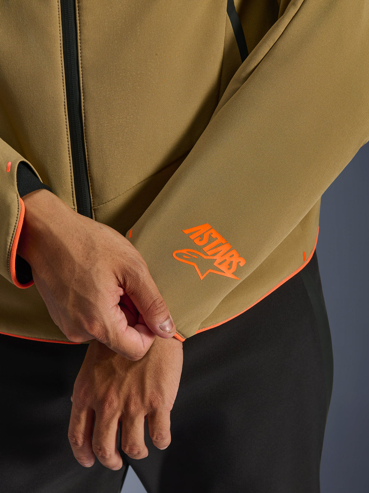 Chaqueta impermeable Alpinestars Aeroshell, color caqui oscuro con detalles en naranja flúor, primer plano del puño de la manga y el dobladillo inferior con el logotipo naranja brillante de Astars, ribetes en naranja neón y detalle de cremallera frontal impermeable, construcción deportiva en softshell.