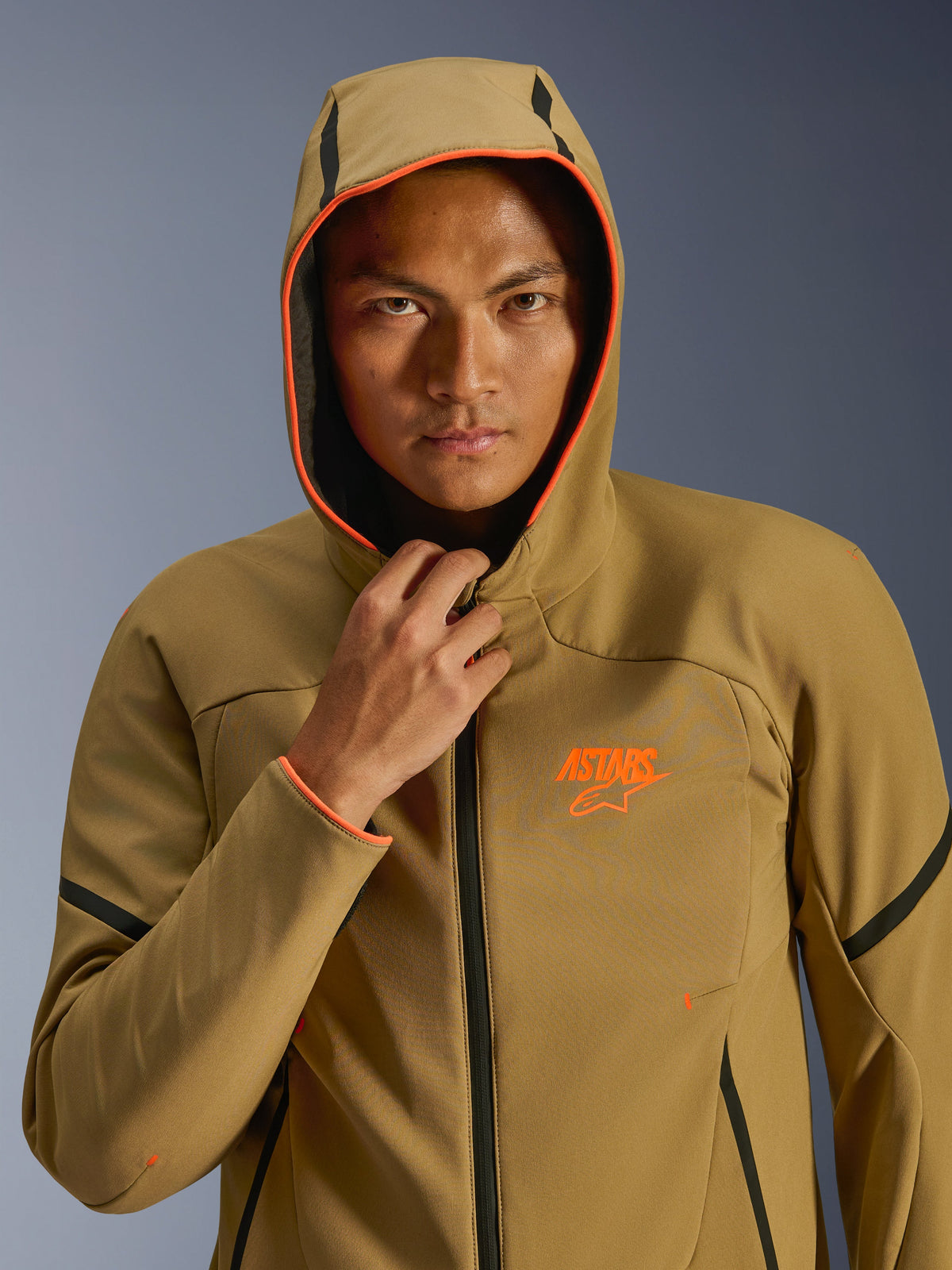 Chaqueta Alpinestars Aeroshell Waterproof, caqui oscuro, negro y naranja flúor, modelo con la chaqueta y la capucha puesta, detalles en naranja flúor vibrante y logotipo ASTARS naranja, elegantes acentos en negro, softshell impermeable de estilo deportivo moderno.