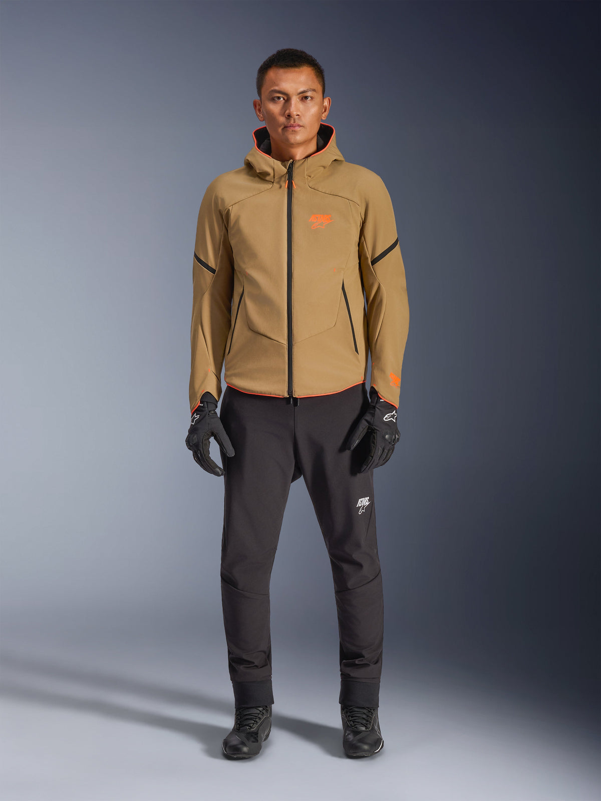 Modelo masculino vistiendo la chaqueta impermeable Alpinestars Aeroshell en color Dark Khaki con detalles en negro y naranja flúor, logo naranja en el pecho y diseño con capucha, combinada con pantalones negros y guantes de moto negros, estilo sport-touring casual
