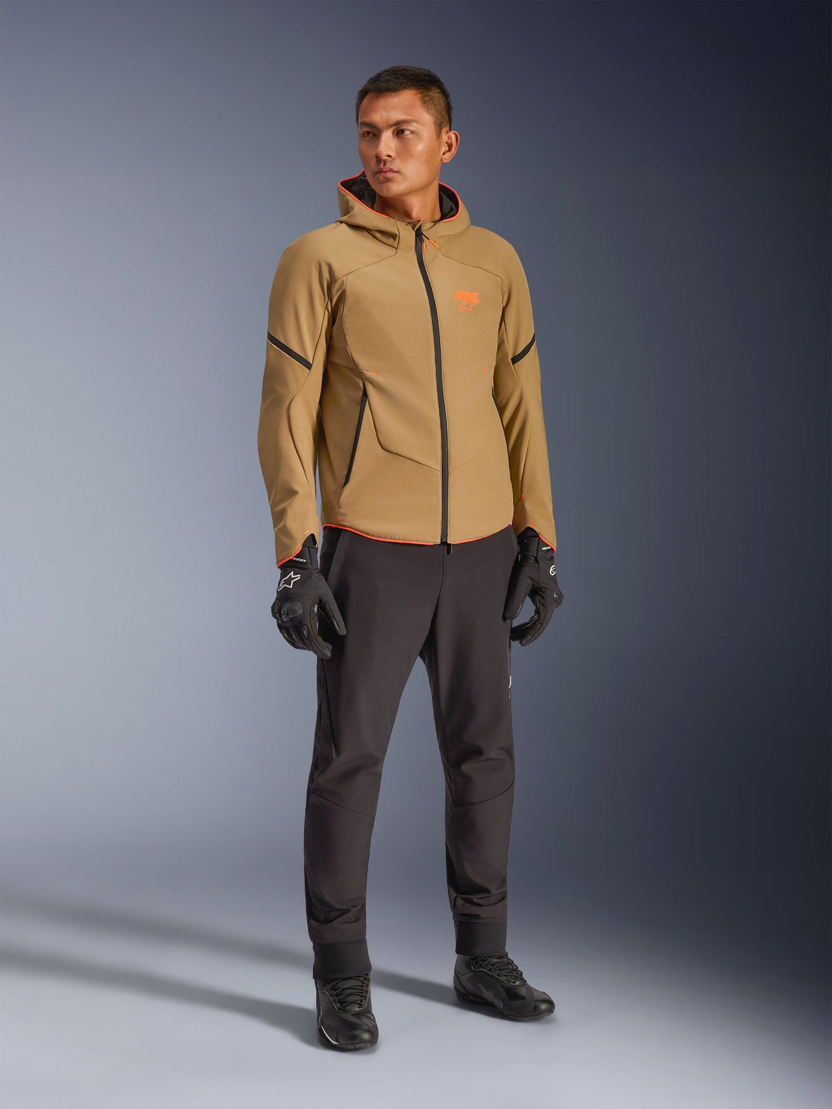 Chaqueta impermeable Alpinestars Aeroshell en caqui oscuro, negro y naranja flúor, usada por un modelo con equipamiento a juego, con un diseño softshell con capucha, cremalleras impermeables y detalles y logotipos en naranja vibrante.