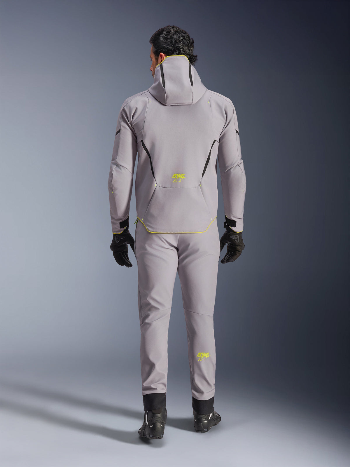 Chaqueta impermeable Alpinestars Aeroshell en Mid Gray Yellow Fluo, vista trasera completa, usada con pantalones grises a juego, guantes negros y calzado de moto negro. Presenta un diseño softshell con capucha con logotipos y detalles 'ASTARS' en amarillo flúor, diseñada para un rendimiento moderno de motociclismo en carretera.
