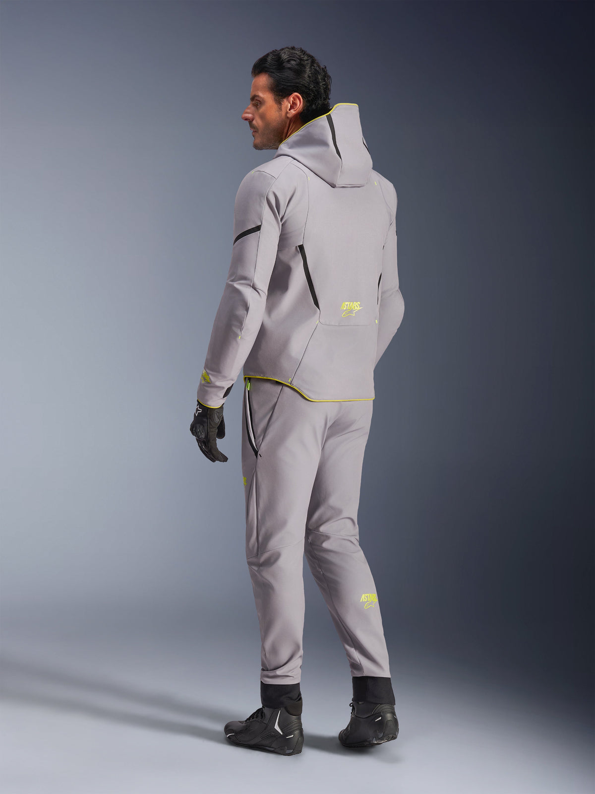 Chaqueta impermeable Alpinestars Aeroshell, gris medio amarillo flúor, usada con pantalones grises a juego, con diseño de sudadera con capucha, detalles y logotipos amarillos, vista trasera, combinada con guantes y botas de conducción negros, ropa de moto moderna.