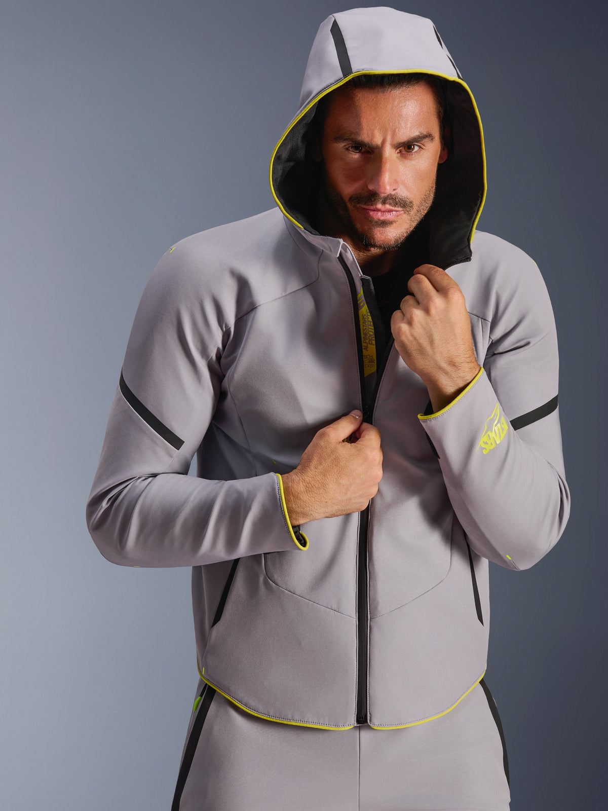 Chaqueta impermeable Alpinestars Aeroshell, gris medio y amarillo flúor, chaqueta softshell deportiva con capucha, lucida por un modelo masculino con la capucha puesta, con cremalleras negras impermeables y detalles en amarillo fluorescente en los puños y el dobladillo, diseño moderno inspirado en el motorsport