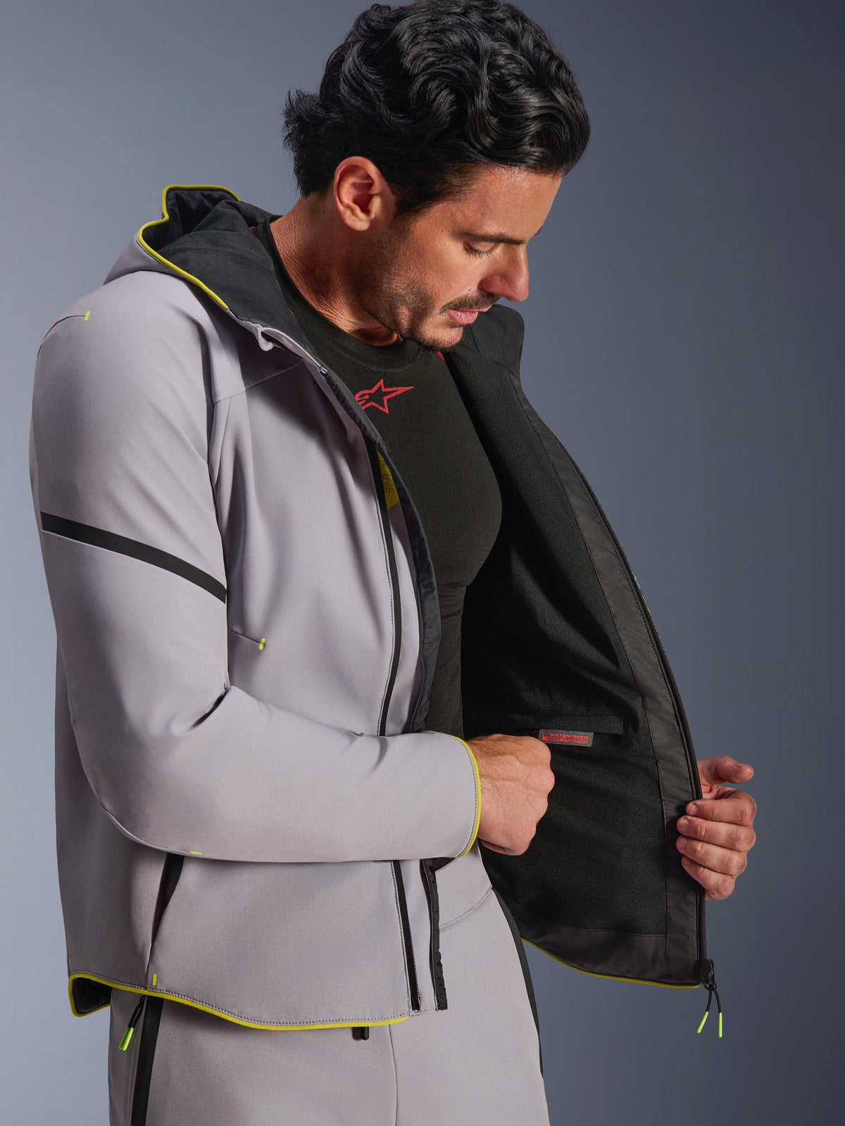Chaqueta impermeable Alpinestars Aeroshell en gris medio y amarillo flúor, mostrada por un modelo masculino con el compartimento impermeable interno y forro negro, sudadera softshell de moto con ribetes amarillos fluorescentes y ajuste atlético moderno.
