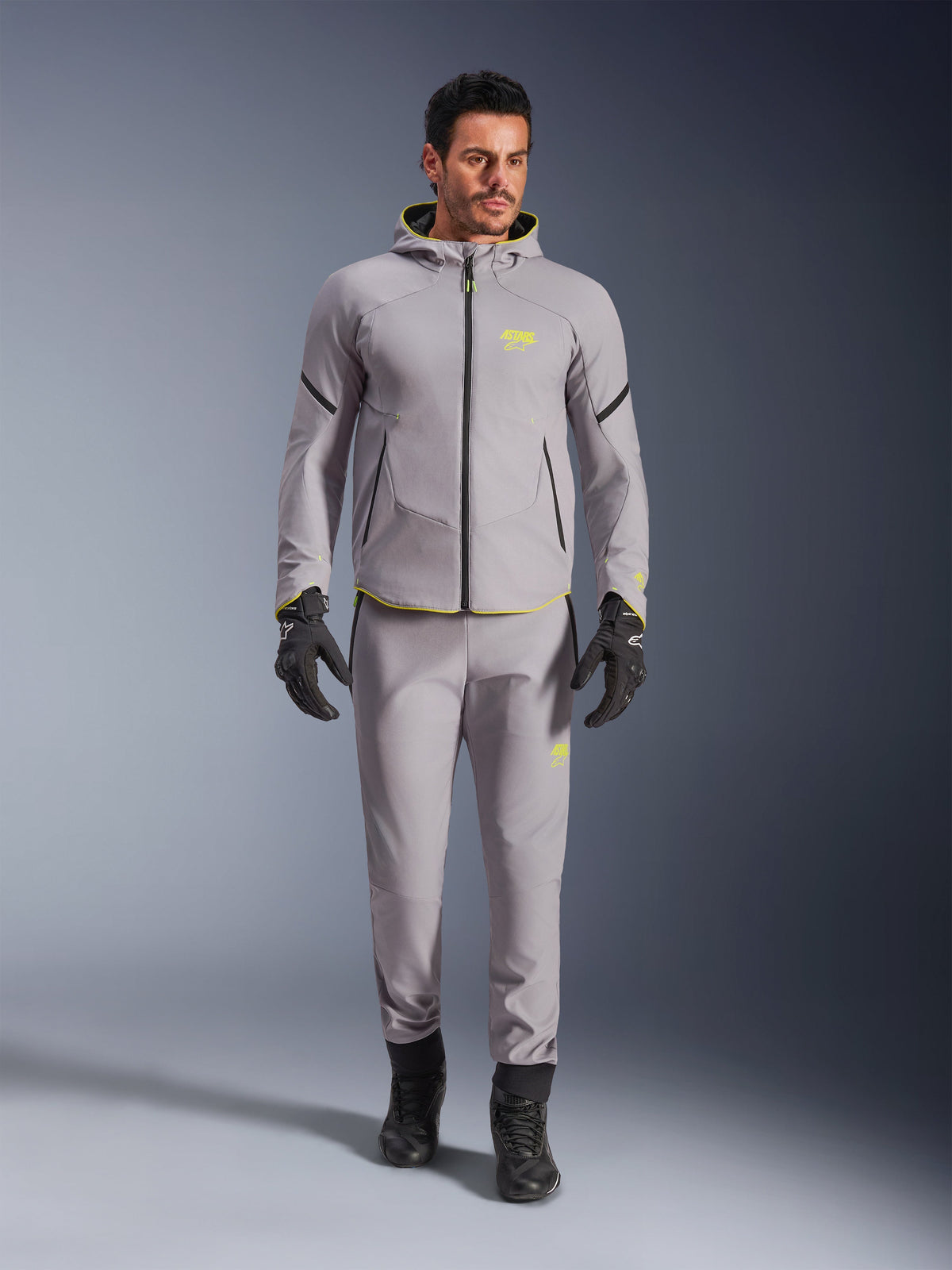 Chaqueta Alpinestars Aeroshell Waterproof, gris medio y amarillo flúor, usada con pantalones grises a juego, guantes y botas negros, con un moderno diseño de sudadera con capucha de estilo deportivo, detalles en amarillo vibrante y ajuste atlético.